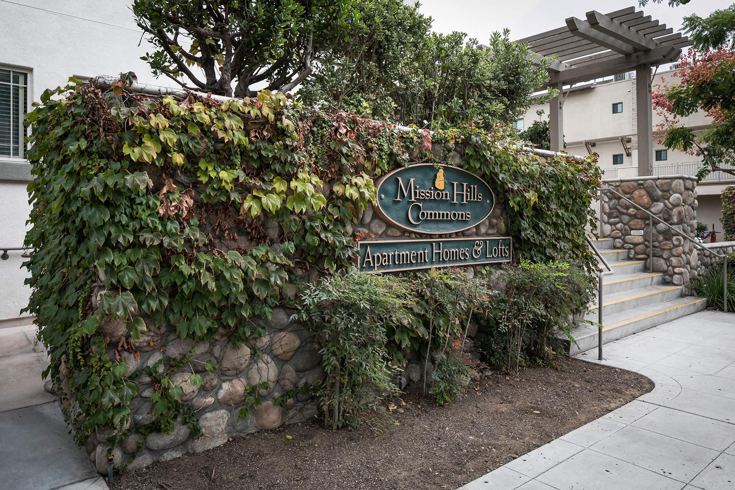 Sign for Mission Hills Commons, featuring lush green ivy and a stone wall. The sign indicates "Apartment Homes & Lots" and is surrounded by landscaped greenery and a staircase leading up to the entrance of the complex.