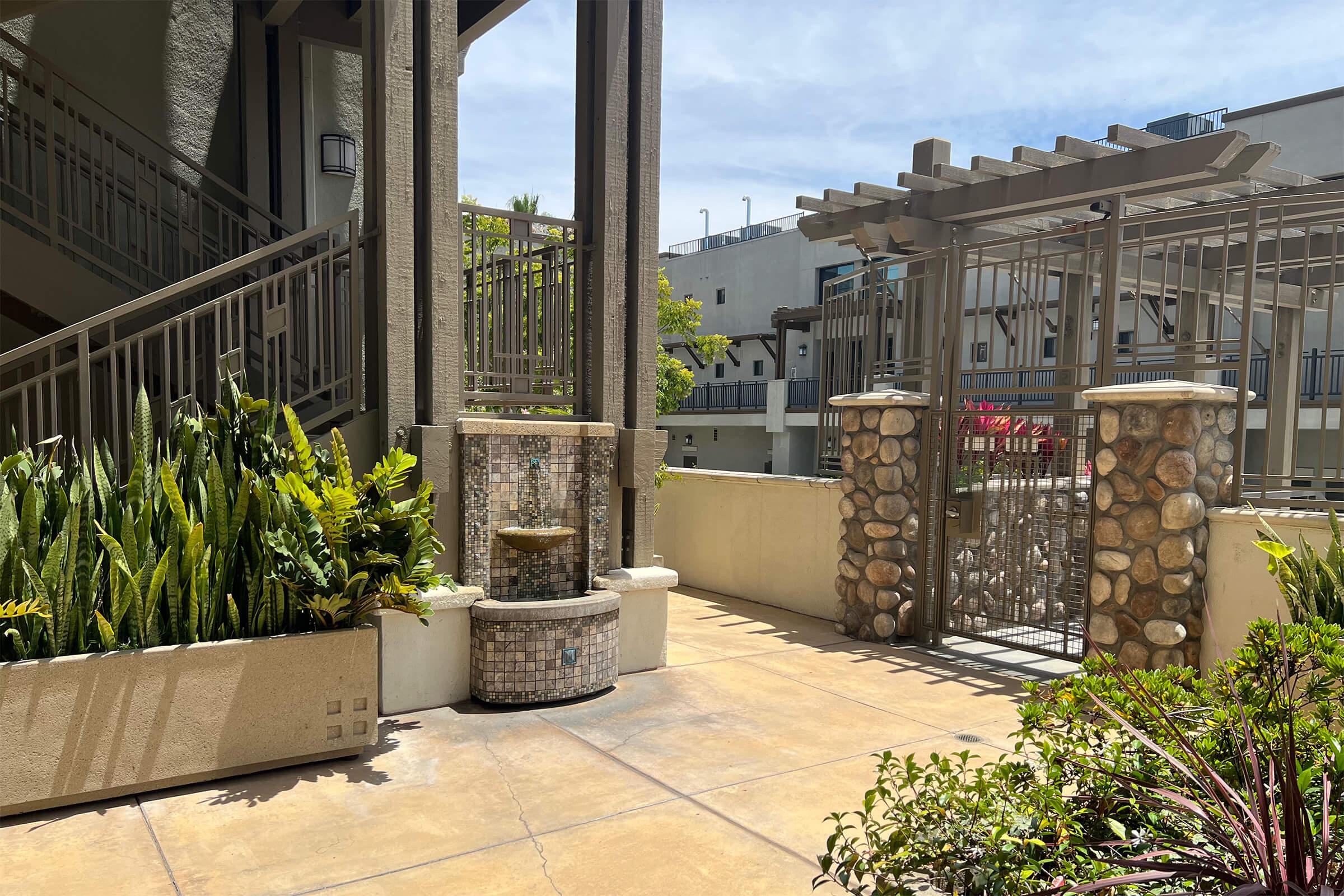 An outdoor area featuring a decorative fountain surrounded by potted plants, stone columns, and a gated entrance. The scene is bathed in natural sunlight, highlighting the textures of the stone and plants, with a background of a building and clear blue sky.