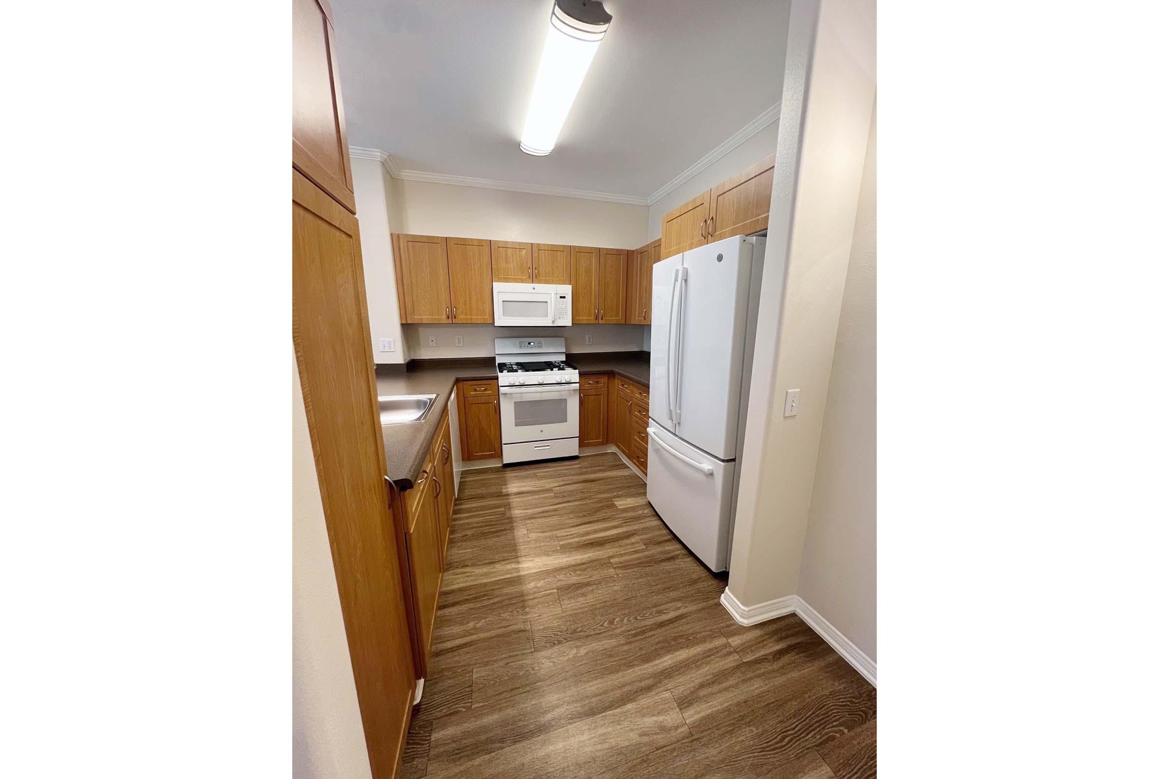 A modern kitchen featuring wooden cabinets, a white refrigerator, and a white stove with an oven. The countertop is gray, and the floor has a wood-like finish. The space is well-lit with overhead lighting, creating a bright atmosphere.