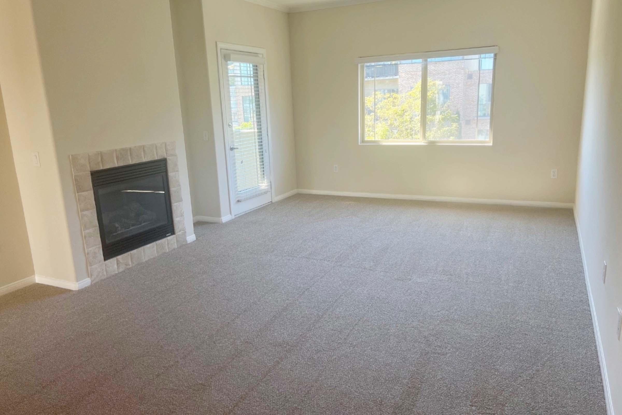 A spacious, empty living room with light beige walls and carpet. Features a stone fireplace on one side and a large window allowing natural light. A door leads to a balcony. The room is well-lit and clean, creating an inviting atmosphere.