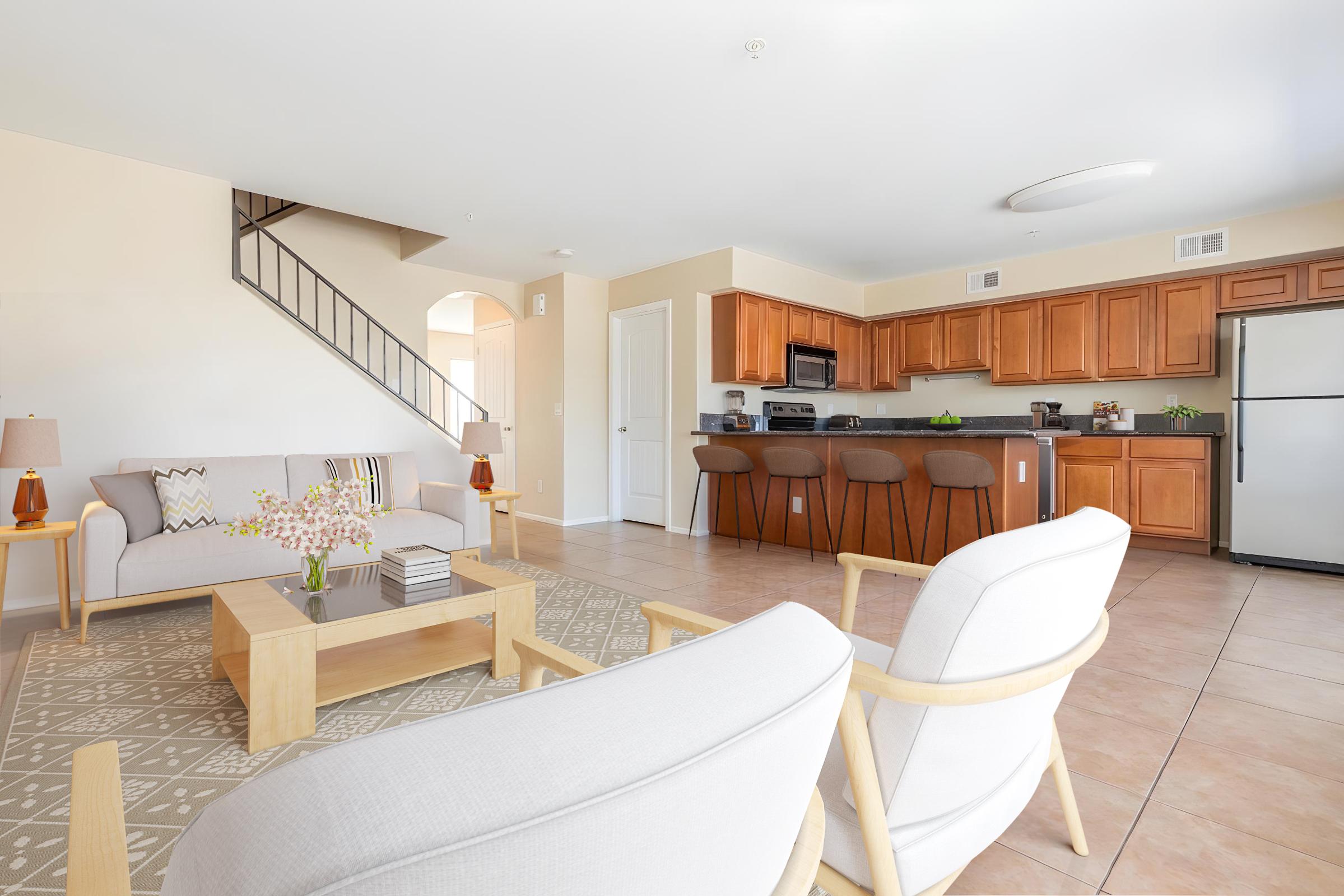 Bright and modern living space featuring a light-colored sofa and armchairs, a wooden coffee table with decorative items, and a kitchen with wooden cabinets and stainless steel appliances. The open layout includes a staircase in the background and a neutral color palette throughout.
