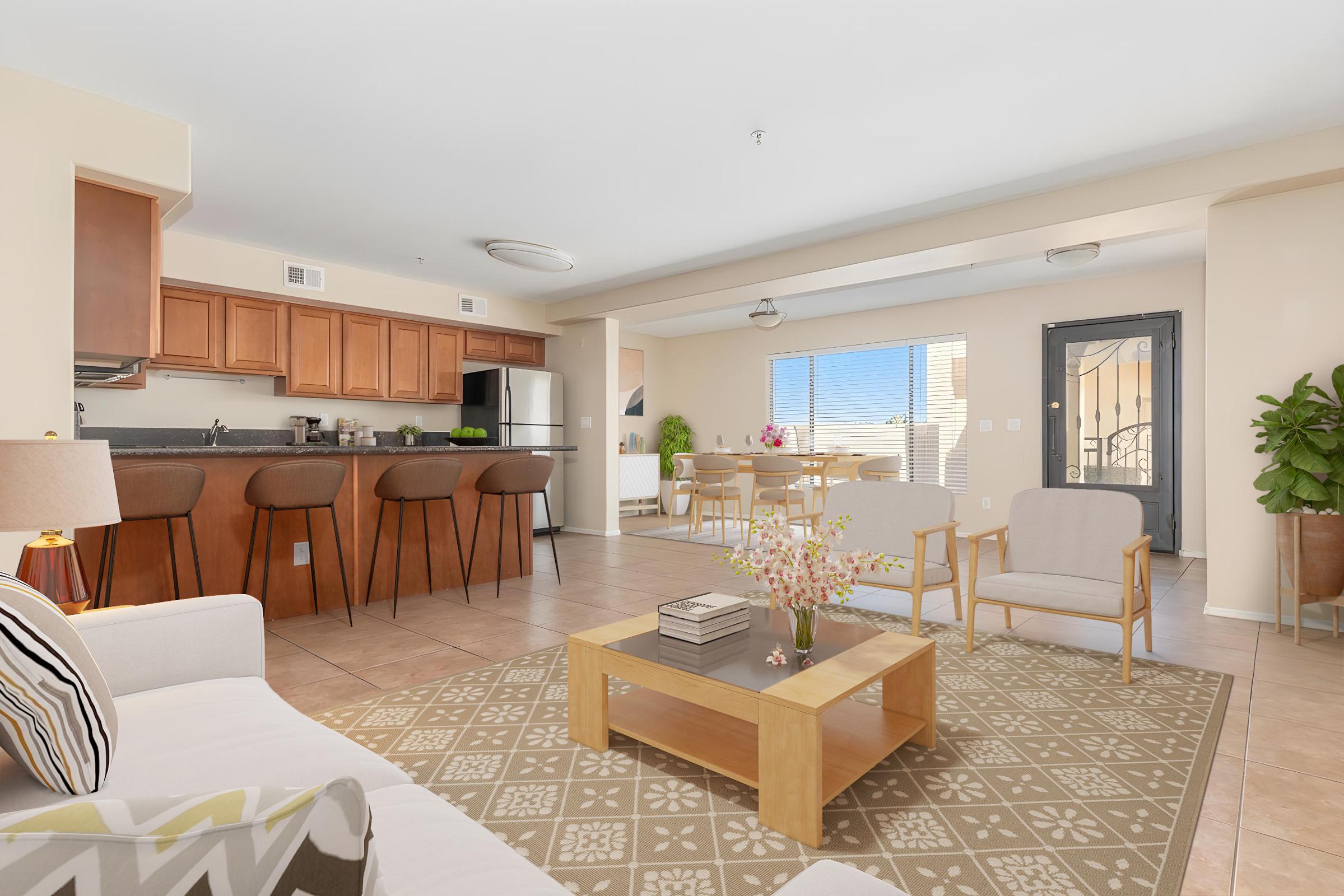 Bright and modern living space featuring a cozy seating area with light-colored furniture, a coffee table, and decorative plants. The open kitchen, with wooden cabinets and bar seating, is visible in the background. Large windows provide natural light, enhancing the inviting atmosphere.