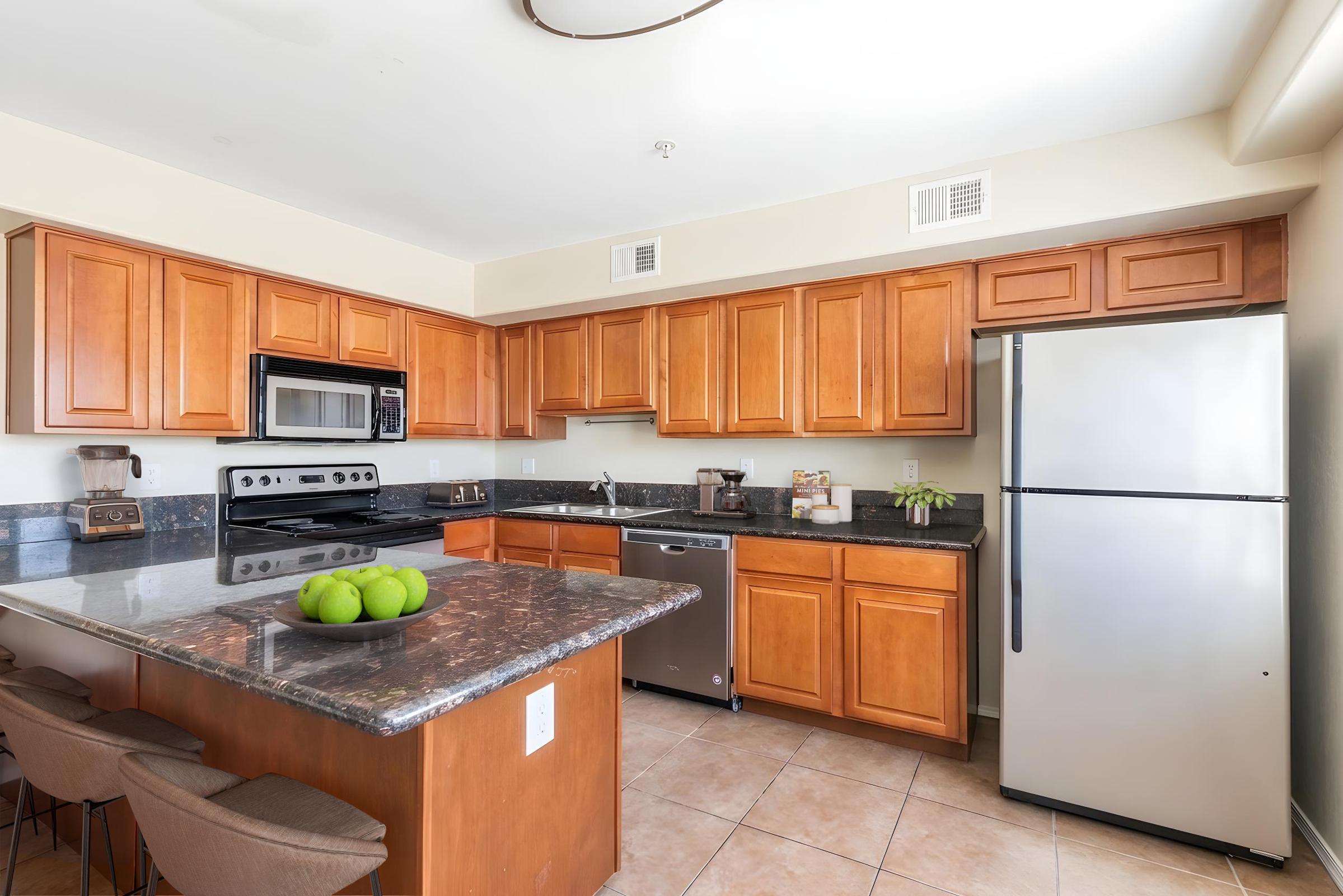 A modern kitchen featuring wooden cabinets, a stainless steel refrigerator, and a black stove with an overhead microwave. The kitchen island has a dark granite countertop and a bowl of green apples. Light-colored tile flooring complements the overall design.