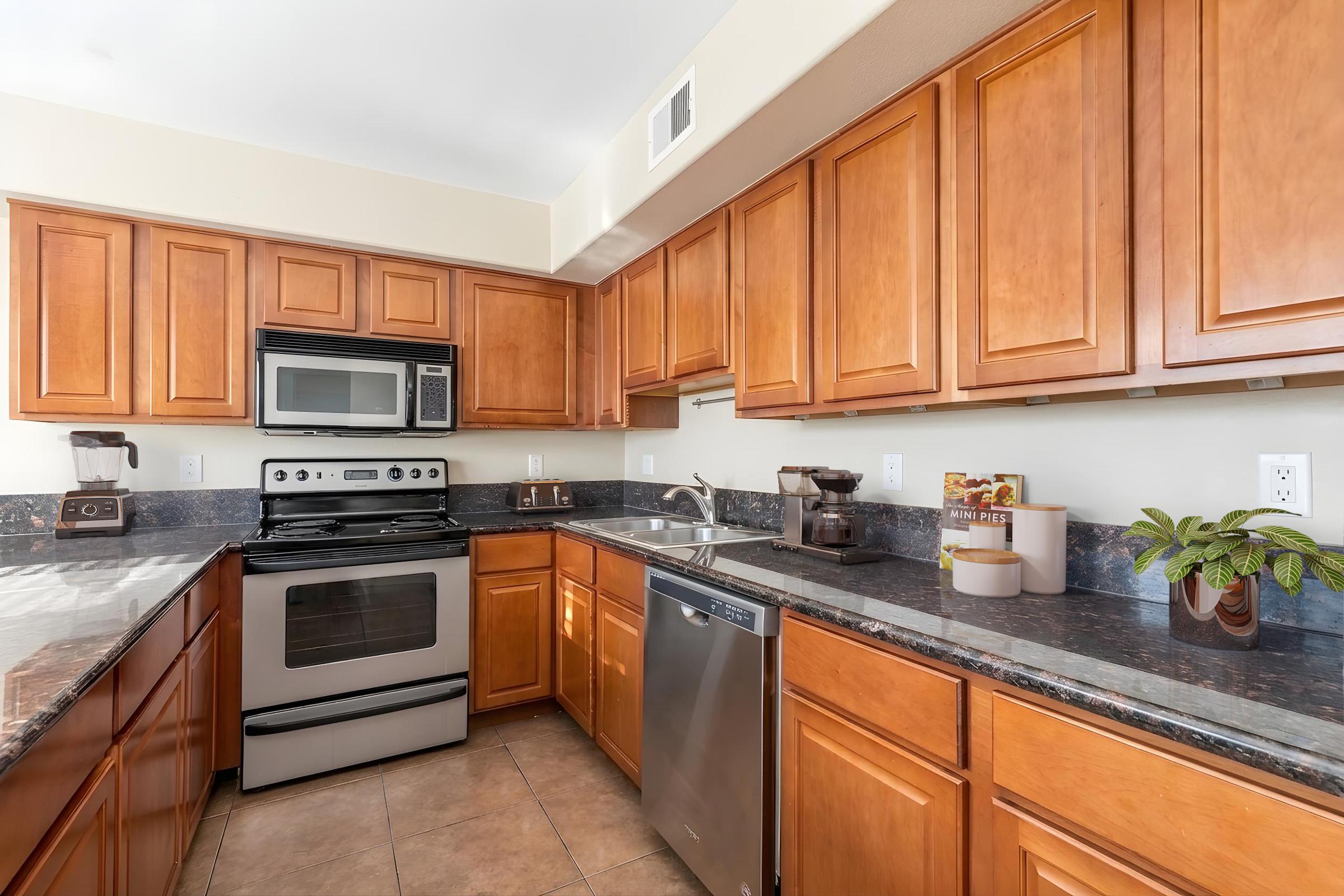 A modern kitchen featuring wooden cabinets, stainless steel appliances including a stove, microwave, and dishwasher, a sink with a faucet, and a countertop with a coffee maker and a small plant. The overall aesthetic is clean and contemporary, with warm tones from the cabinetry and granite surfaces.