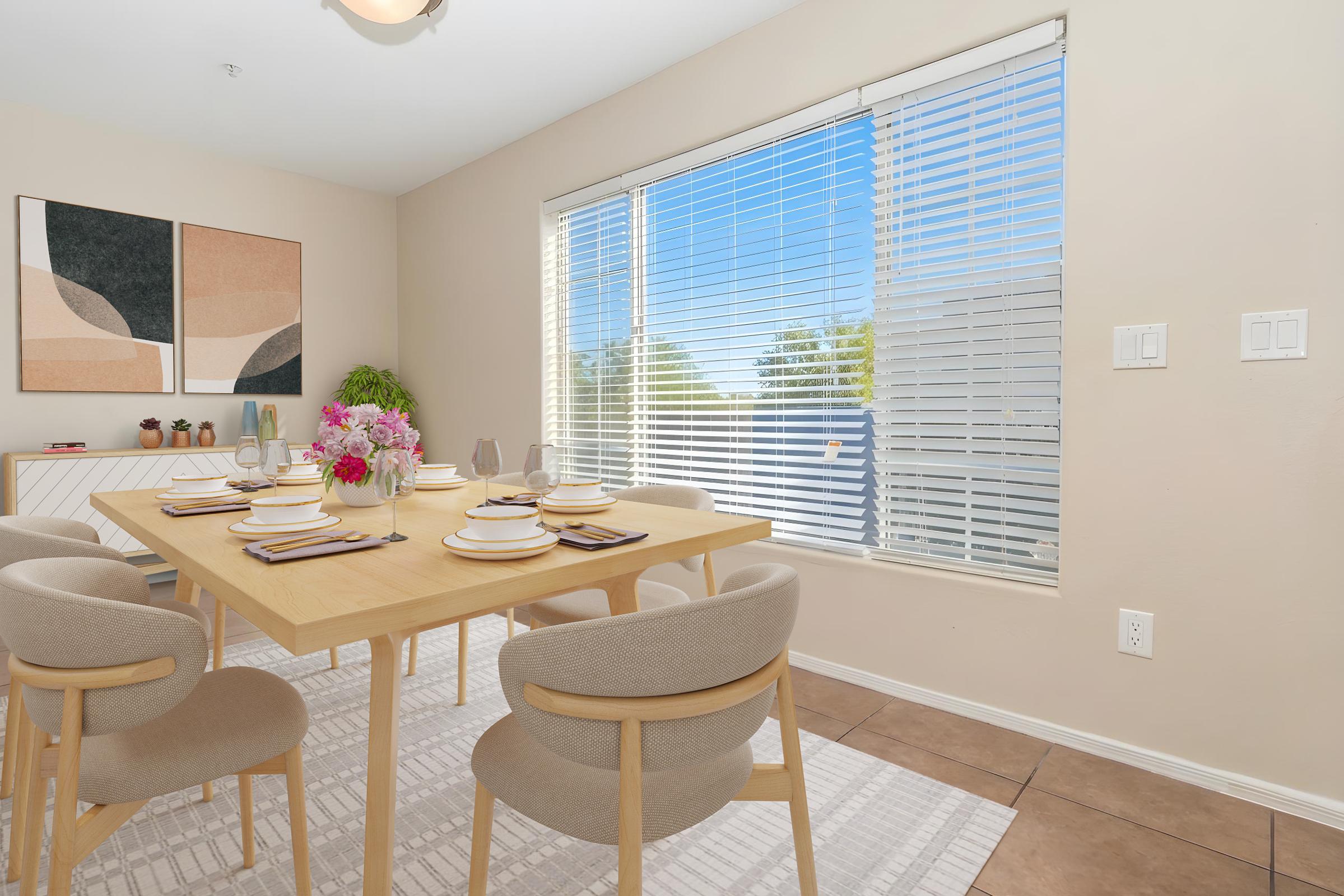 A bright dining area featuring a light wooden table set for six with dishes and glasses. Surrounding the table are light-colored chairs. A vase of flowers adds a pop of color. The room has large windows with blinds that let in natural light, and there are abstract art pieces on the wall, creating a modern, inviting atmosphere.