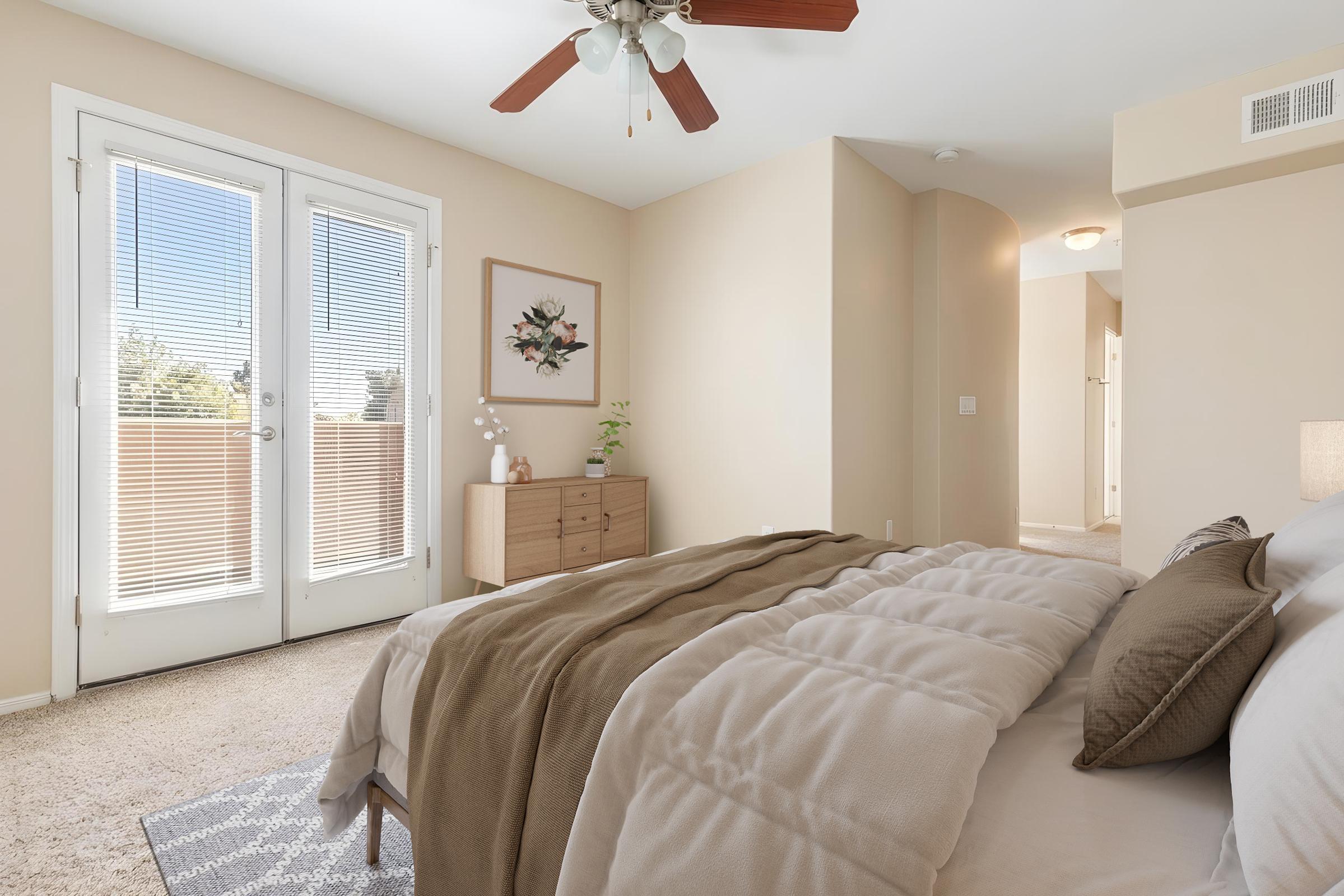 A cozy bedroom featuring a queen-sized bed with a neutral-colored comforter and decorative pillows. There’s a dresser against the wall, a potted plant on top, and large double doors leading to a balcony. The walls are a soft beige, and natural light fills the room from the open doors.