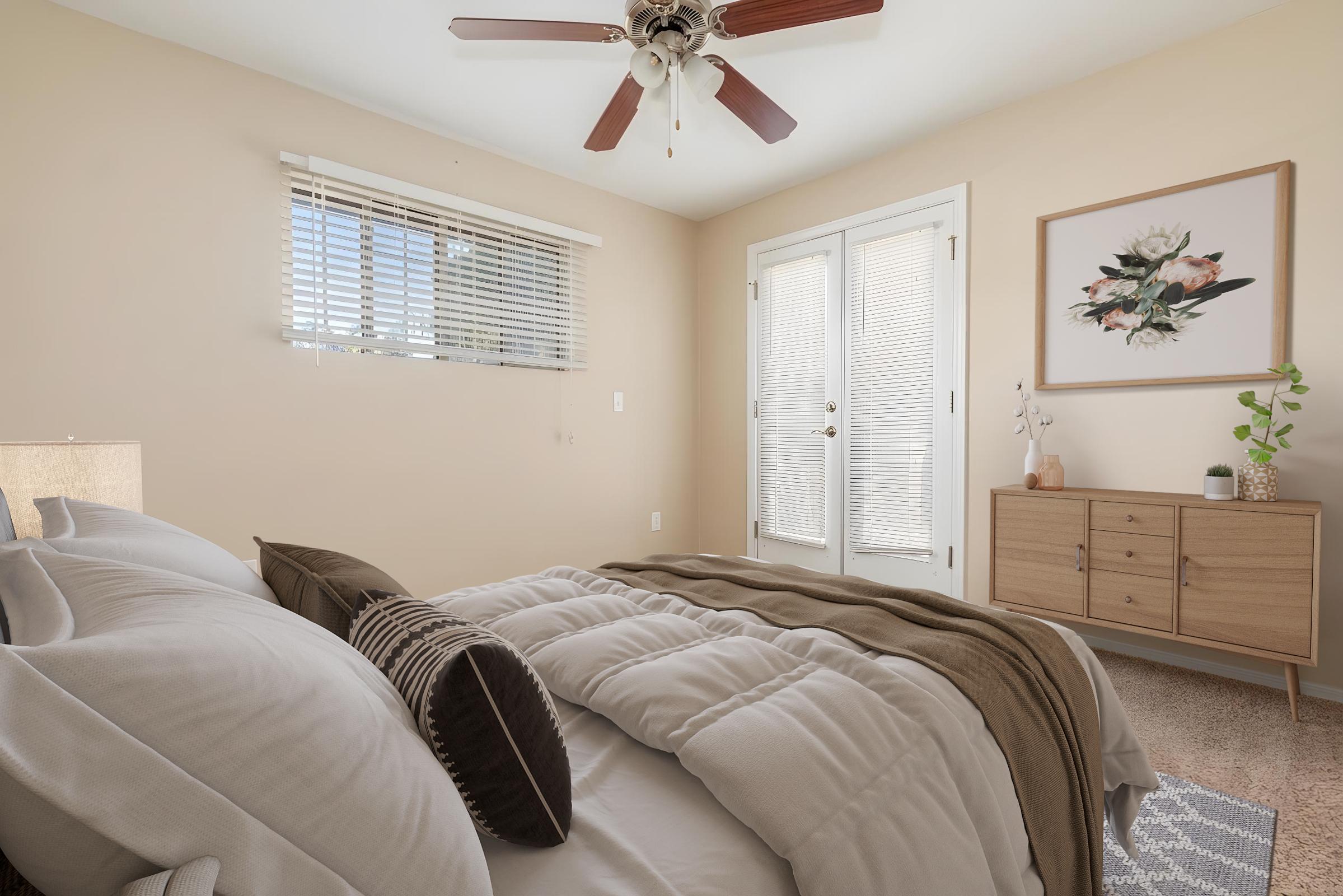 A cozy bedroom featuring a neatly made bed with decorative pillows, a wooden nightstand with a lamp, and a framed floral artwork on the wall. Natural light filters through white blinds, creating a warm ambiance. A potted plant adds a touch of greenery, while double doors provide access to a balcony or outdoor area.