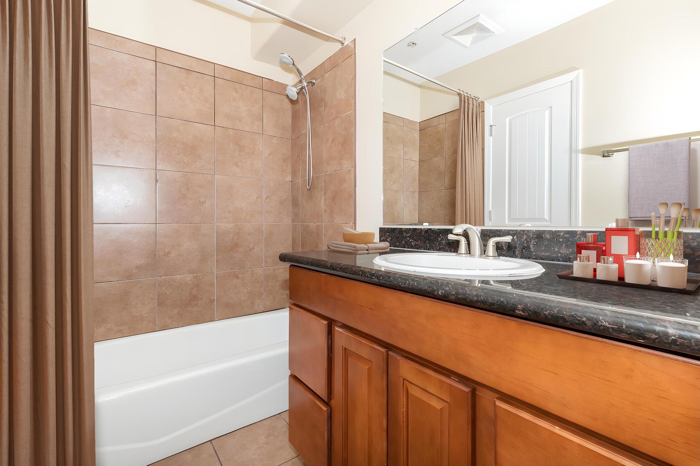 A clean bathroom featuring a bathtub with a showerhead, tiled walls in warm tones, a wooden vanity with a black countertop, and a mirror. Decorative items like candles and toiletries are arranged neatly on the counter, and there's a shower curtain on the side. The overall ambiance is modern and inviting.
