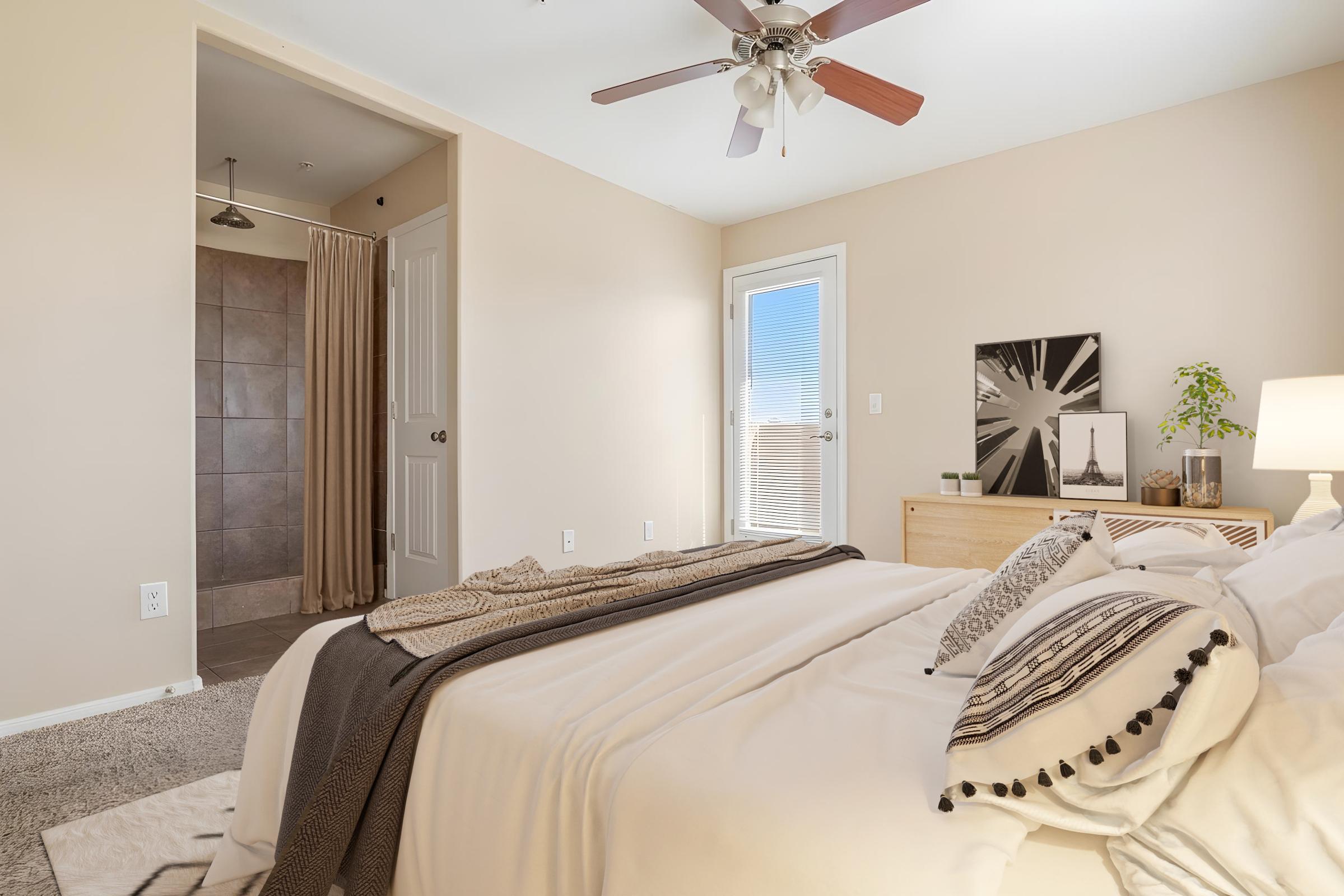 A cozy bedroom with a neatly made bed featuring decorative pillows and a throw blanket. A nightstand displays a small plant and artwork. A bathroom area, with a shower curtain, is visible in the background. Natural light streams in through a window, enhancing the warm tones of the room.