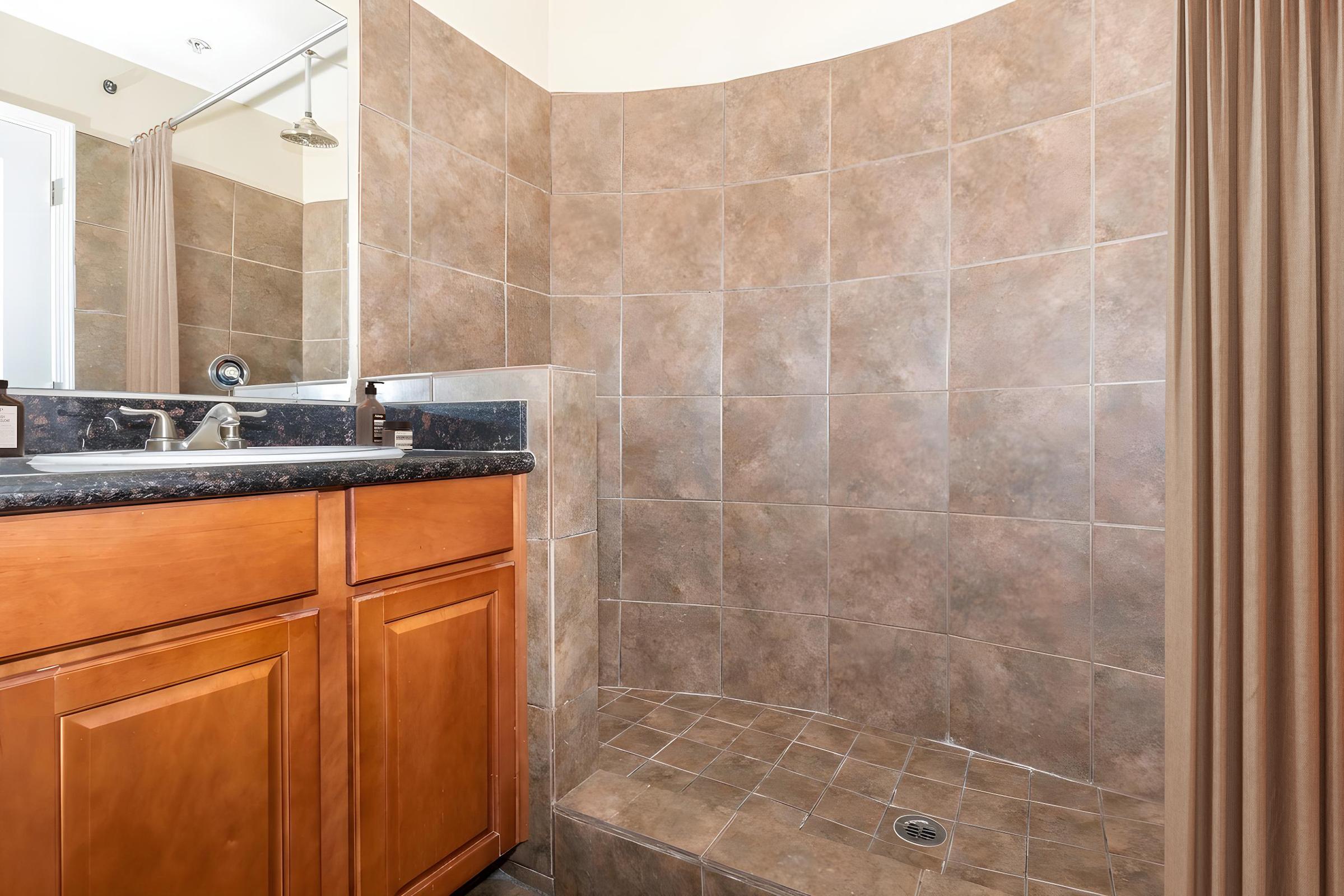 A modern bathroom featuring a curved tile shower area with brown tiles, a granite countertop on a wooden vanity with drawers, and a mirror above. A showerhead is mounted on the wall, and nearby is a cream-colored curtain draping over the shower entrance.