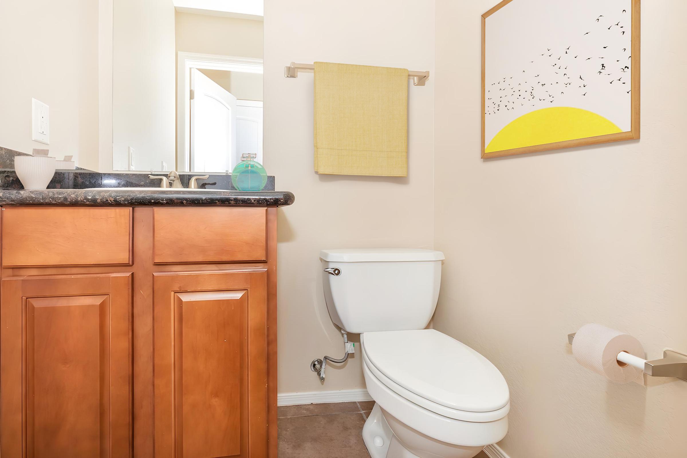 A small bathroom featuring beige walls, a dark granite countertop with a sink, and wooden cabinetry. There is a white toilet, a yellow towel hanging on a rack, and a framed artwork depicting a sun and birds. A pale pink toilet paper roll is on a holder nearby. The room is well-lit and inviting.