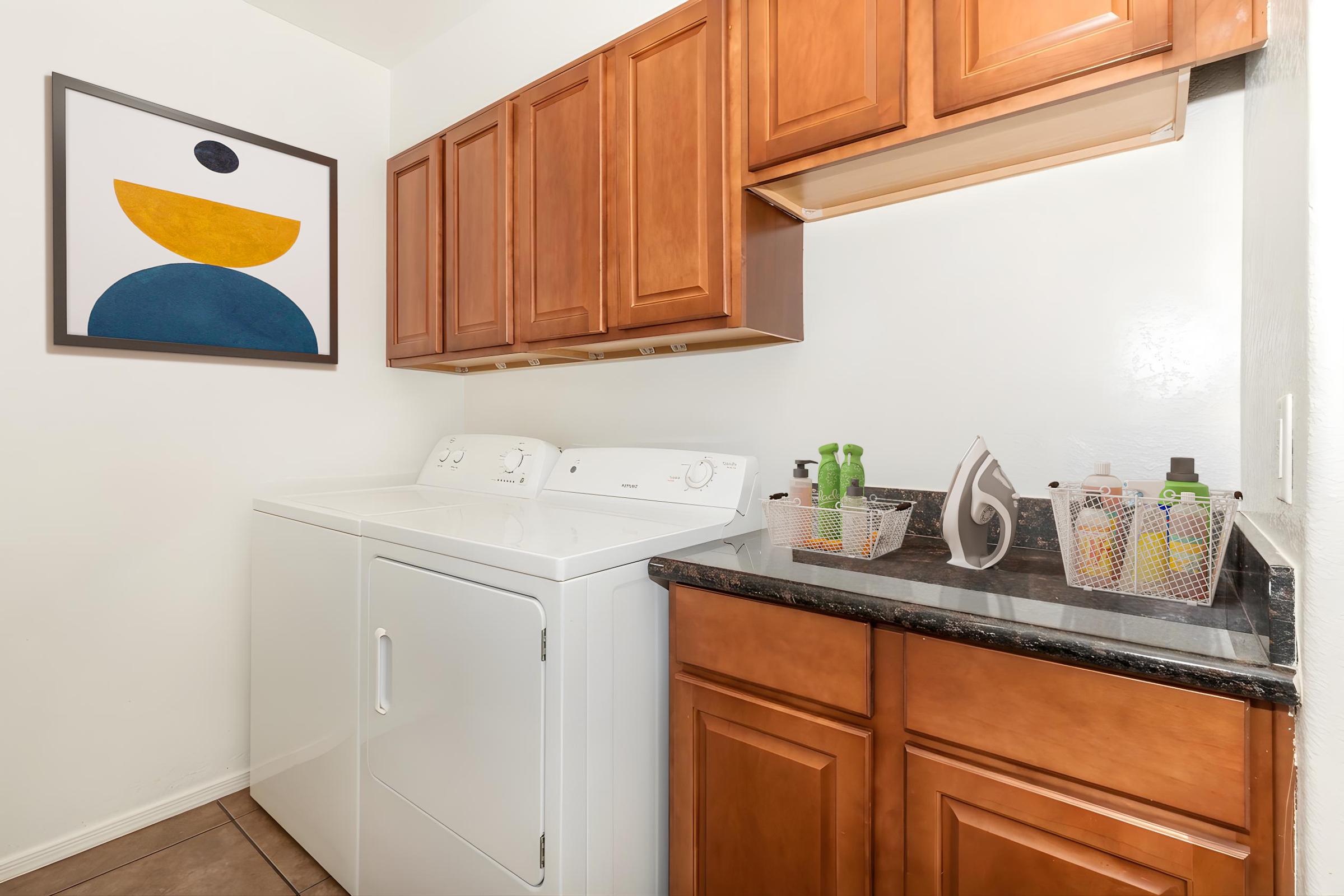 A cozy laundry room featuring a white washing machine and dryer stacked side by side. Above the appliances is a brown wooden cabinet, and a countertop holds various laundry supplies in baskets. A colorful abstract artwork is displayed on the wall, adding a modern touch to the space.