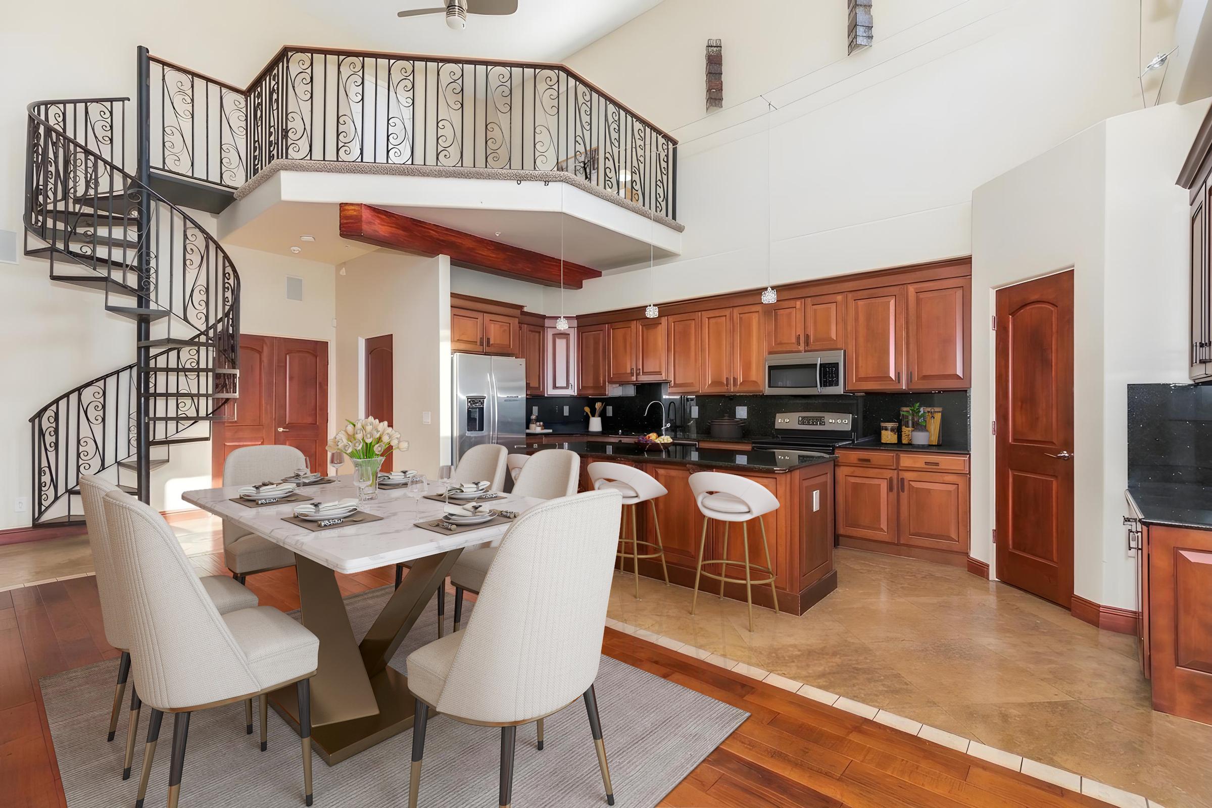 A modern dining area featuring a stylish table set for six, surrounded by elegant upholstered chairs. The room showcases warm wooden cabinetry and a spiral staircase leading to a balcony. The kitchen is equipped with stainless steel appliances, and large windows allow ample natural light.