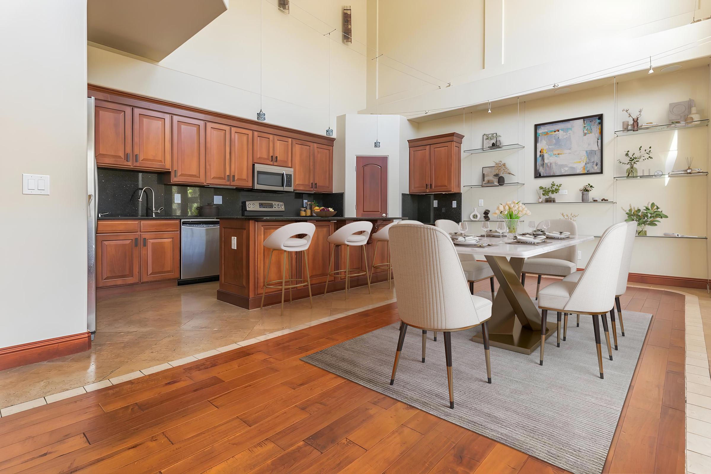 A modern kitchen and dining area featuring wooden cabinetry, a black countertop, and stainless steel appliances. A stylish dining table with six chairs is set on a patterned rug, and the space is adorned with decorative shelves showcasing plants and artwork, illuminated by natural light from large windows.