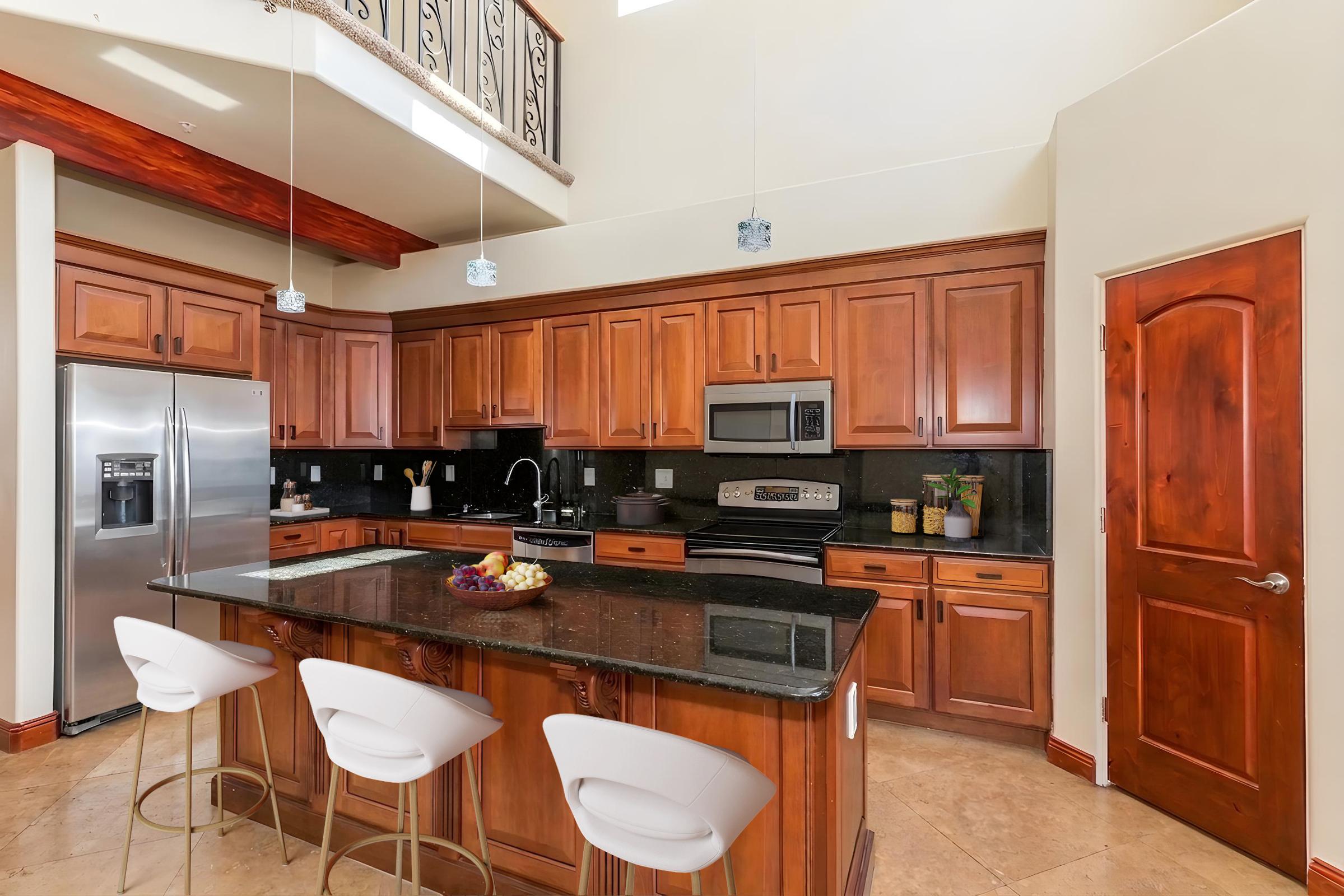 Spacious modern kitchen featuring dark wooden cabinetry, stainless steel appliances, and a large central island with seating. The countertops are black, and there is natural light from windows above. Decor includes potted plants and a bowl of fruit on the island. The flooring is light-colored tile.