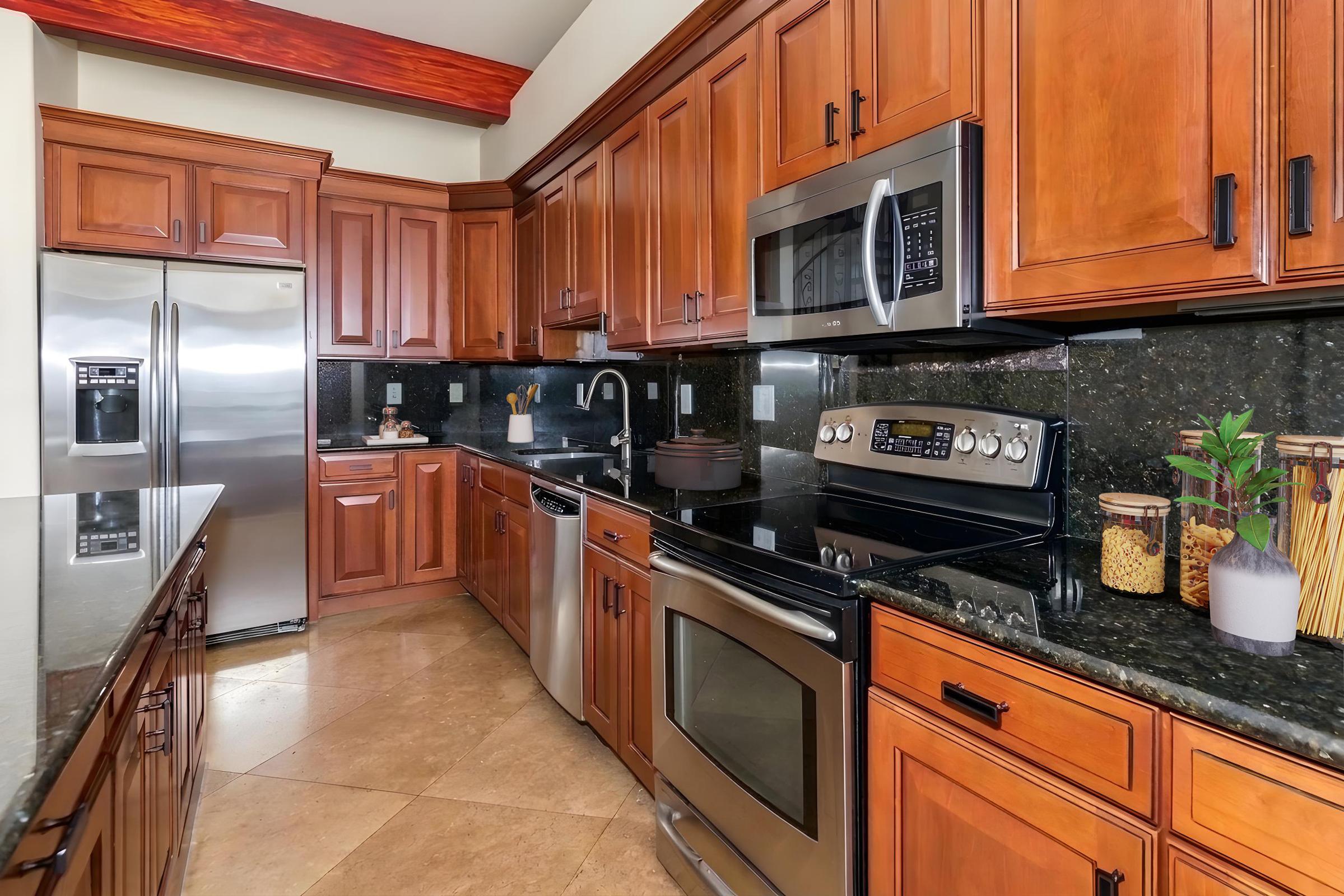 Modern kitchen featuring wooden cabinetry, stainless steel appliances, and dark granite countertops. It includes a refrigerator, microwave, oven, and dishwasher. A small plant and jars with utensils are visible on the counter. The kitchen is well-lit with a warm wooden ceiling.
