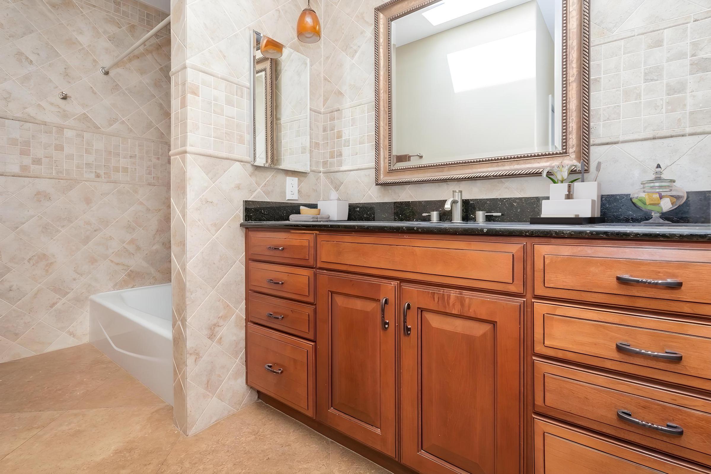 A modern bathroom featuring a wooden vanity with multiple drawers and a black countertop. Above the vanity is a decorative mirror. On the left side, a bathtub is visible, alongside a tiled shower area with beige tiles. Natural light illuminates the space from a ceiling skylight.