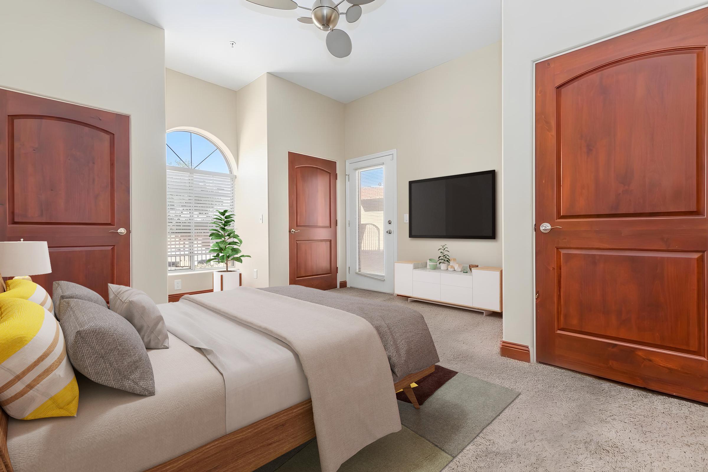 A cozy bedroom featuring a neatly made bed with gray and yellow pillows, a light brown bed frame, and a soft beige throw. The room has large wooden doors, a flat-screen TV mounted on the wall, and a small plant beside a white media console. Natural light filters in through a window, creating a warm atmosphere.