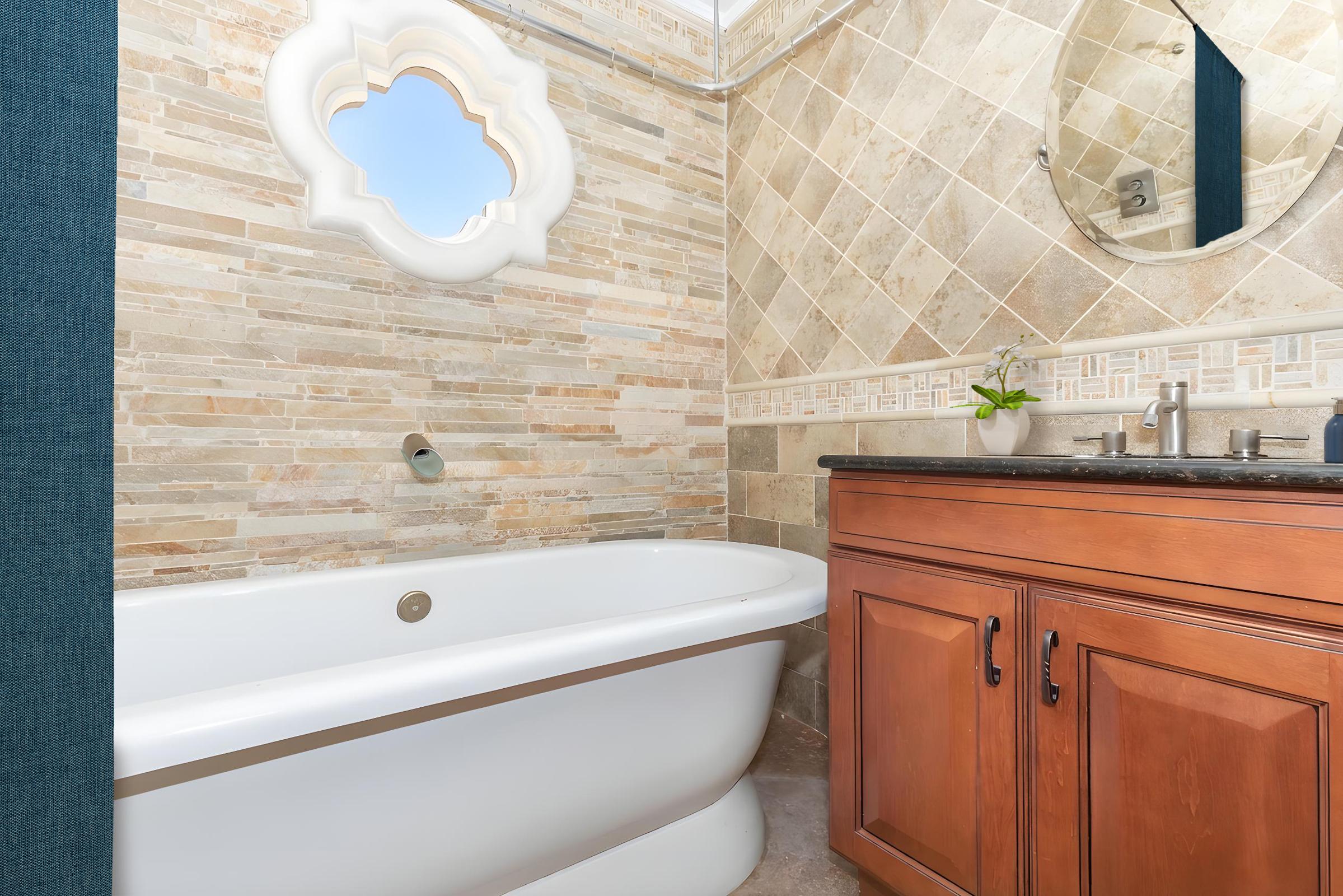A beautifully designed bathroom featuring a freestanding bathtub, wooden cabinetry, and a round mirror. The walls are adorned with textured stone tiles in neutral tones, and a unique window design adds a decorative element. A small plant is placed on the countertop for a touch of greenery.