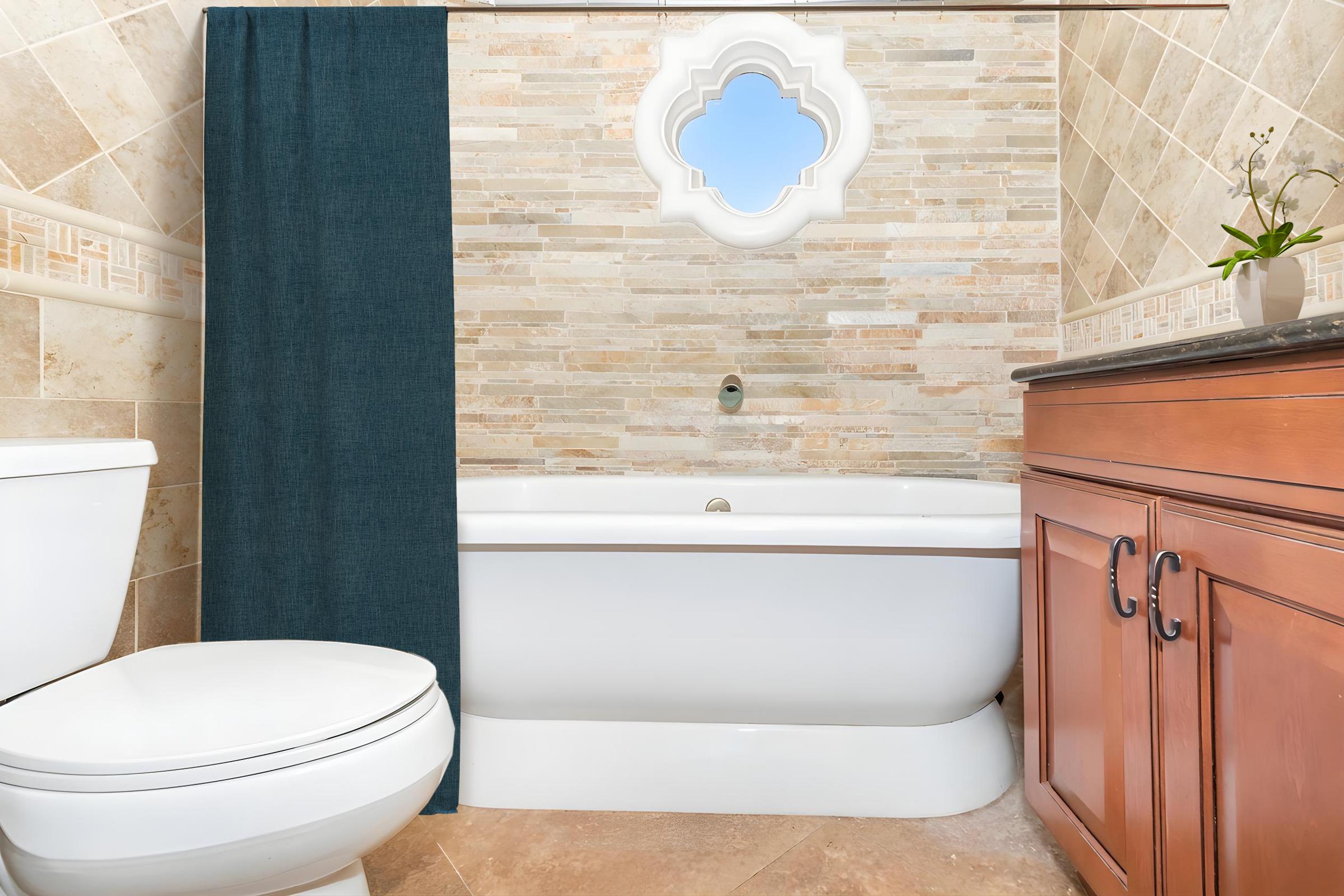A well-lit bathroom featuring a freestanding white bathtub next to a wooden cabinet with drawers. A blue curtain hangs over a window with a decorative arch, and a small plant is placed on the cabinet. A modern toilet is positioned nearby on the tiled floor.