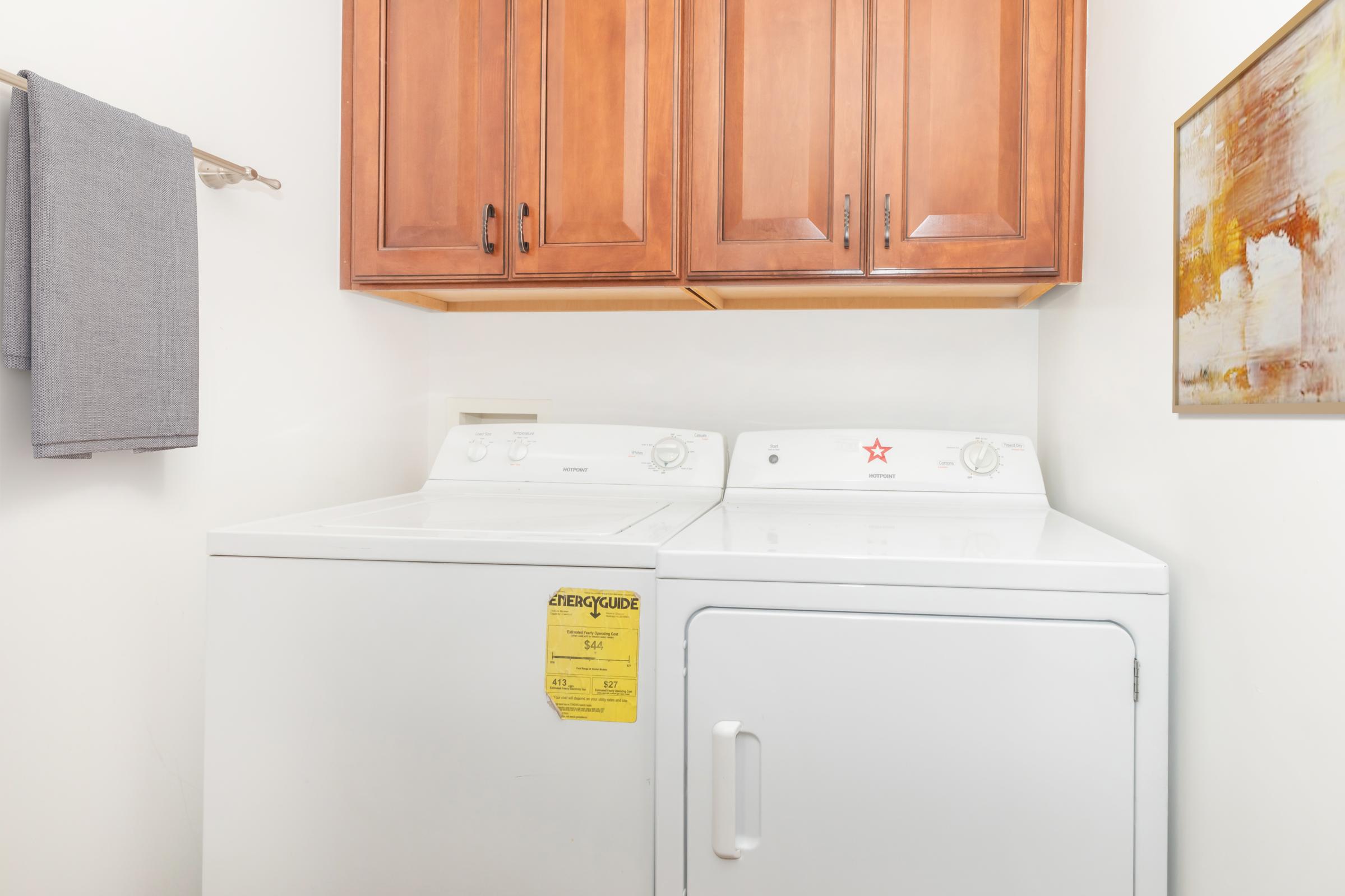 A laundry room featuring a white washer and dryer stacked side by side. Above the appliances are wooden cabinets with a warm finish. A gray towel hangs on a wall-mounted rack nearby, and a colorful abstract painting is displayed on the adjacent wall.
