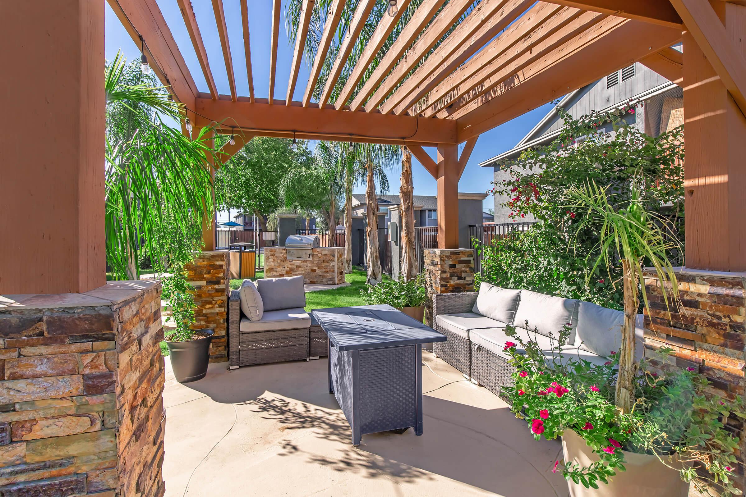 A cozy outdoor seating area under a wooden pergola, featuring comfortable gray sofas with cushions, a small table, and potted plants with colorful flowers. Lush greenery surrounds the space, creating a relaxing atmosphere in a backyard setting under clear blue skies.