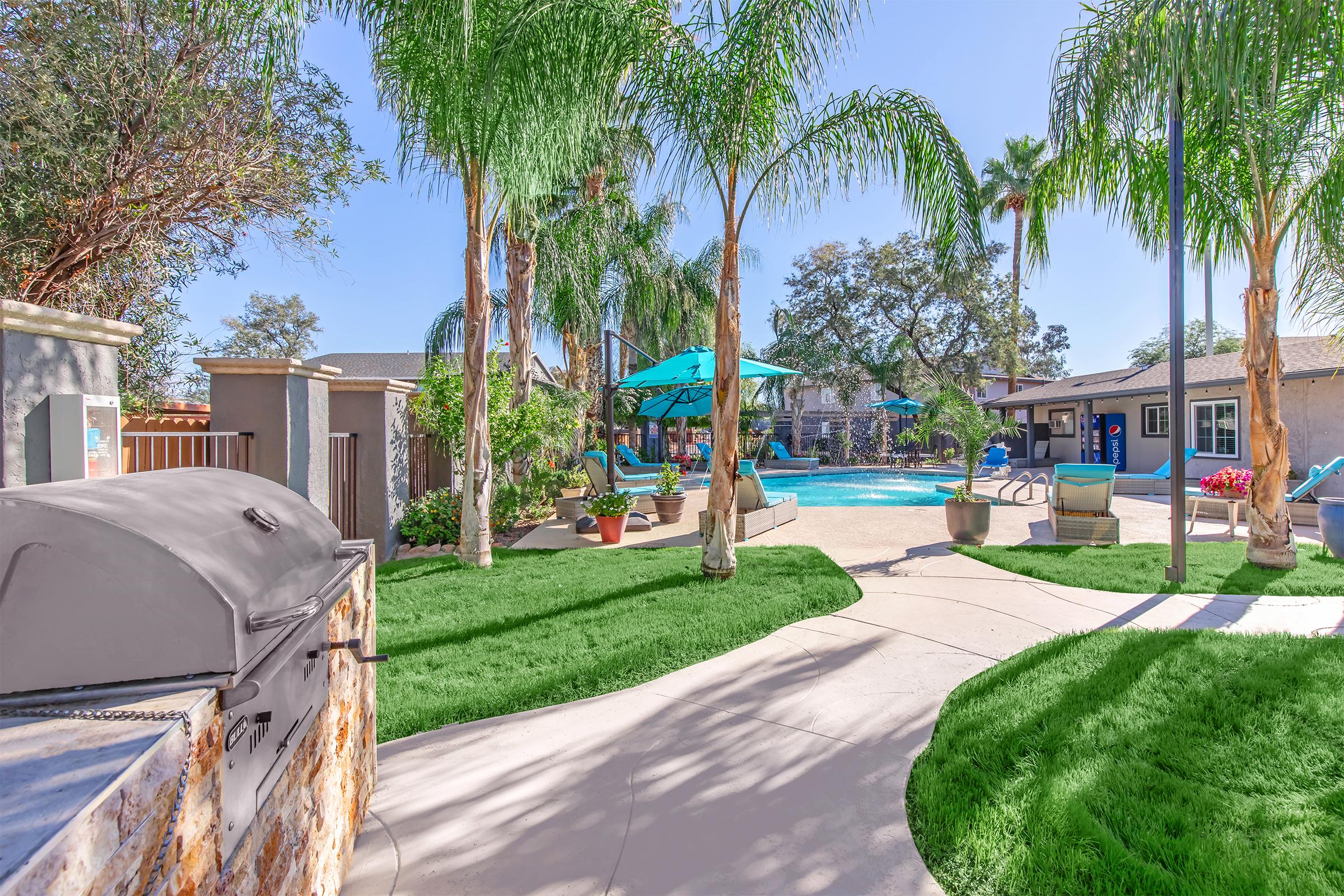 A sunny pool area surrounded by palm trees and lush green grass. In the foreground, there is a grill, and in the background, a sparkling swimming pool is visible with lounge chairs and umbrellas. Flowers are in pots, and there are pathways leading around the space, creating a welcoming outdoor atmosphere.