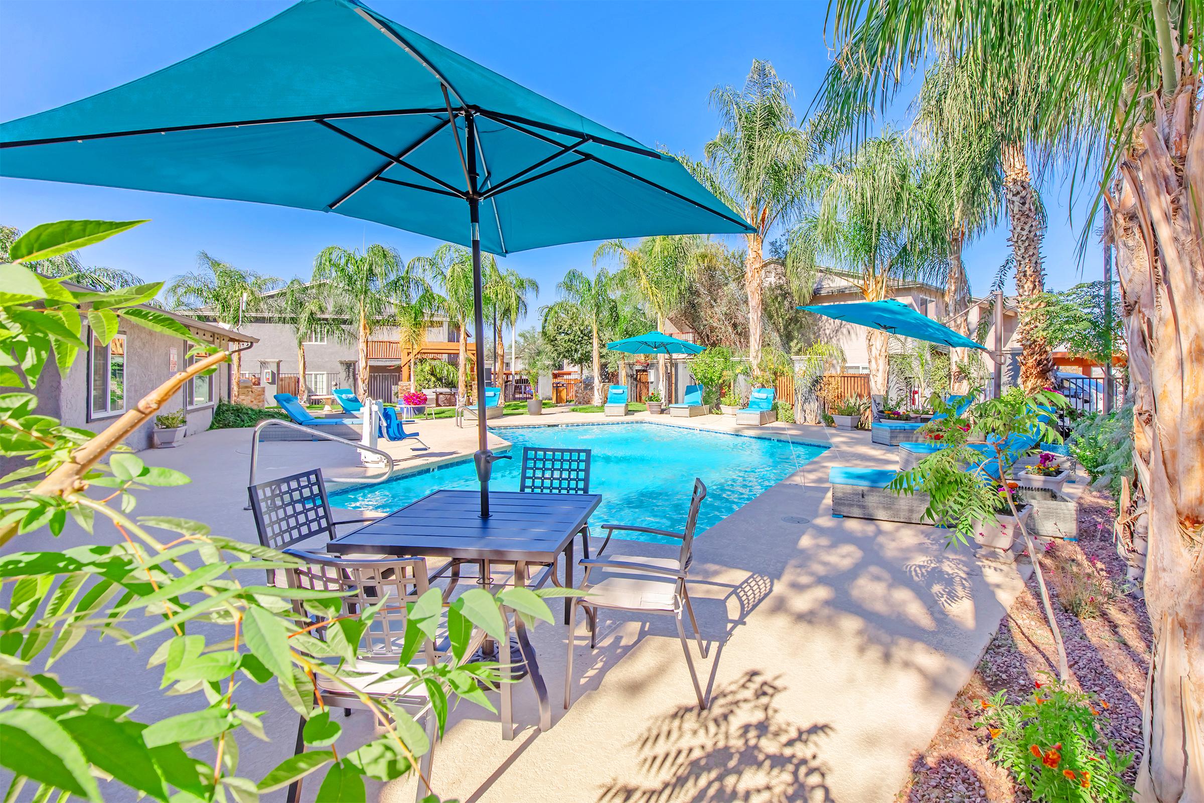 A vibrant outdoor pool area featuring a crystal-clear swimming pool surrounded by palm trees, lounge chairs, and umbrellas. A dining table with chairs sits nearby, creating a relaxing atmosphere perfect for leisure and enjoyment under a bright blue sky.