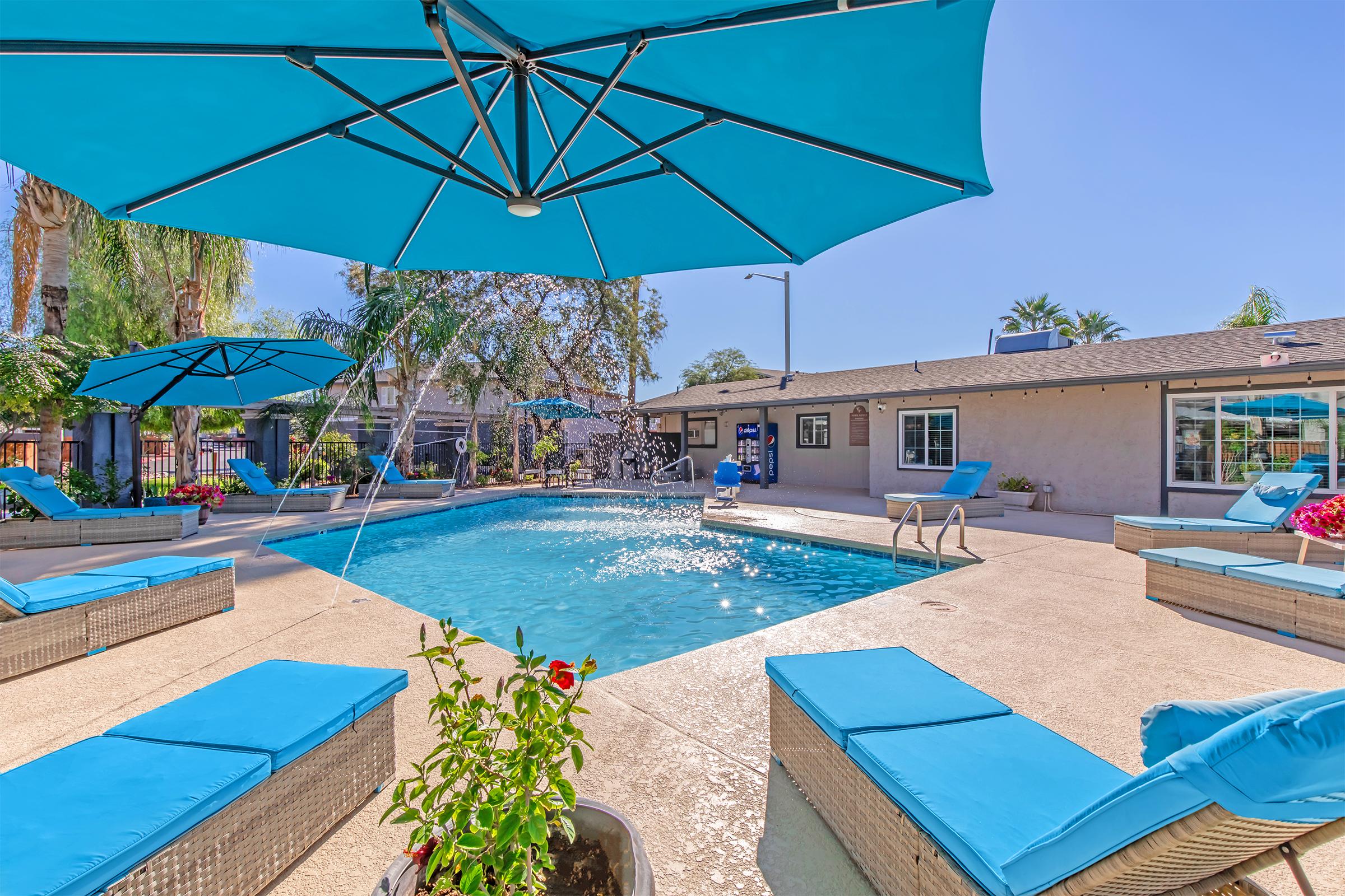 A bright outdoor pool area featuring a clear blue pool, surrounded by lounge chairs with turquoise cushions and large umbrellas. Lush greenery and flowers enhance the setting, while a small fountain adds a touch of elegance. A cozy house is visible in the background under a sunny blue sky.