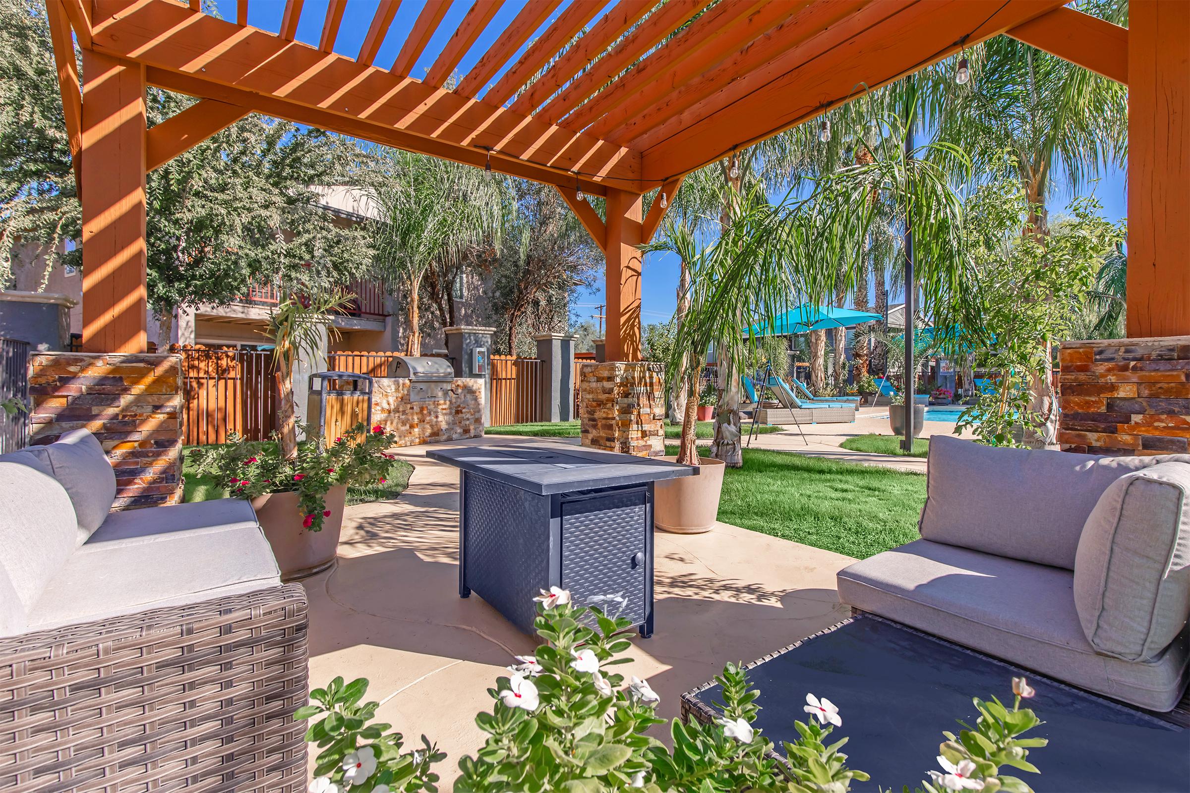 A cozy outdoor seating area under a wooden pergola, featuring two cushioned lounge chairs and a central fire pit table. Surrounding plants add greenery, and in the background, swimming pool and lounge chairs are visible, creating a welcoming, relaxing atmosphere.