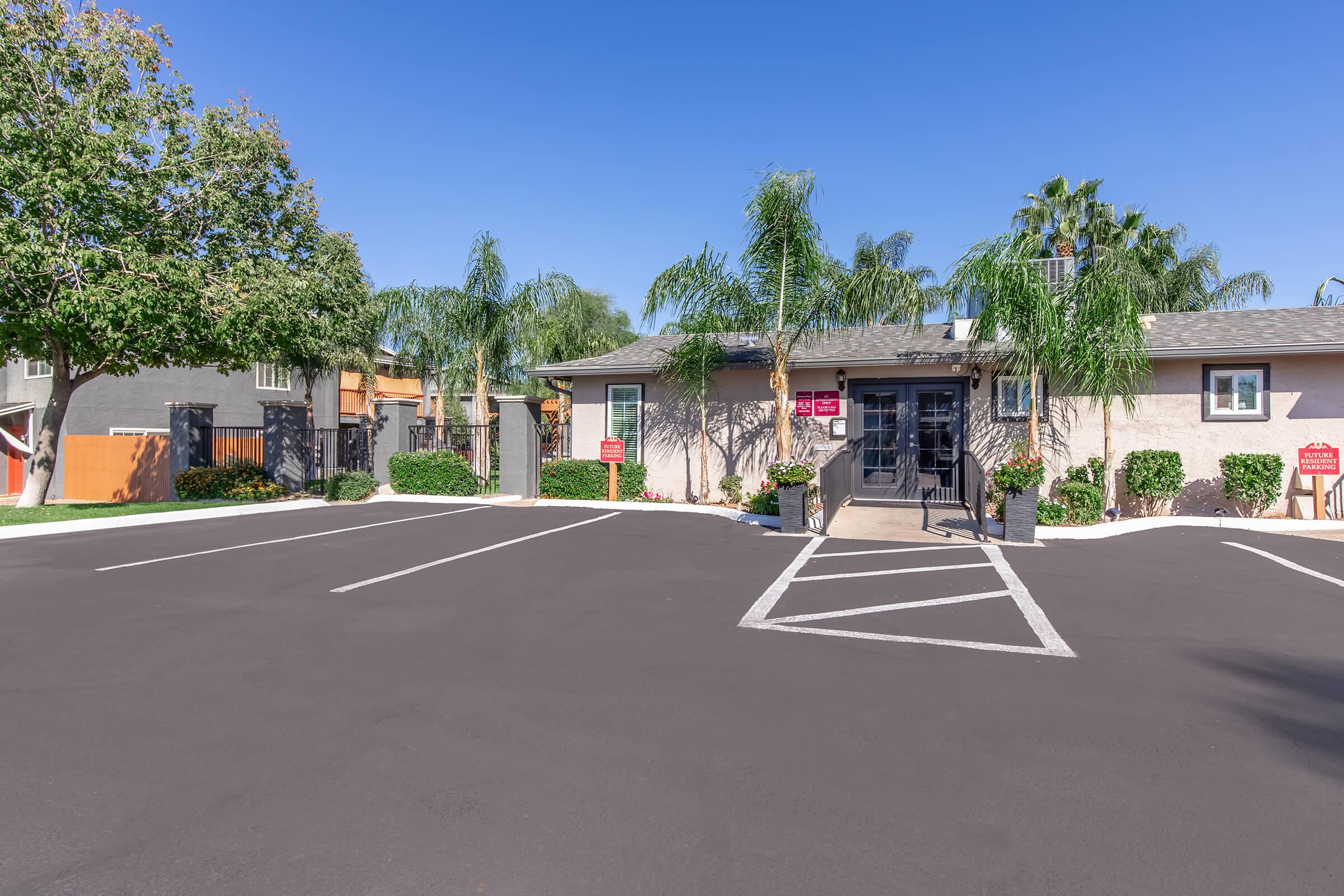 A residential building with palm trees in front, featuring a welcoming entrance. The parking lot shows clearly marked spaces. The surrounding area is well-maintained, with greenery and a clear blue sky overhead. There are signs indicating property management or services at the entrance.