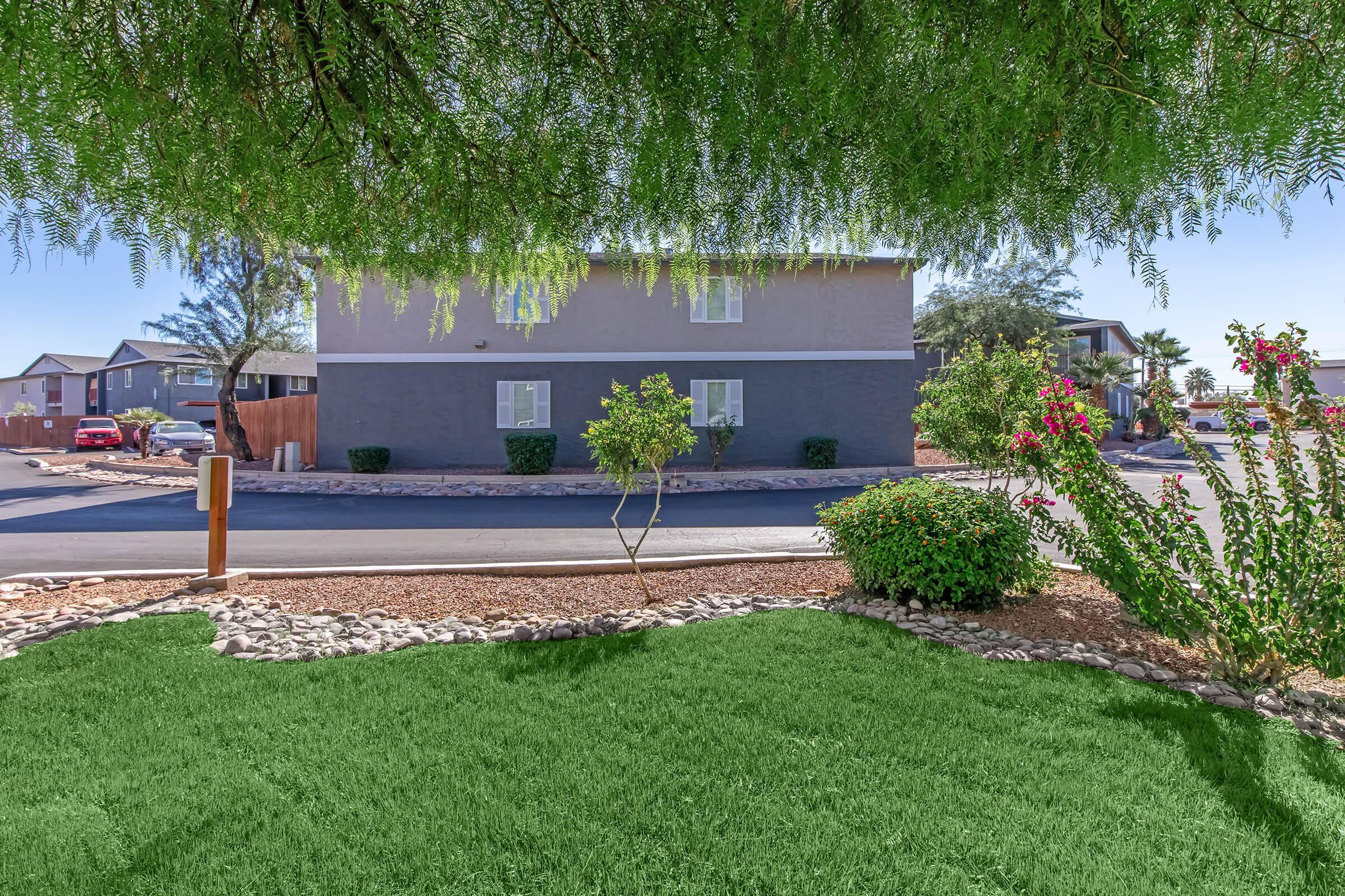 A view of a landscaped area featuring green grass and shrubs, with a multi-story building in the background. The scene is partly shaded by a tree, and there are flowerbeds along the pathway leading to the building. Several vehicles are parked nearby in the background.