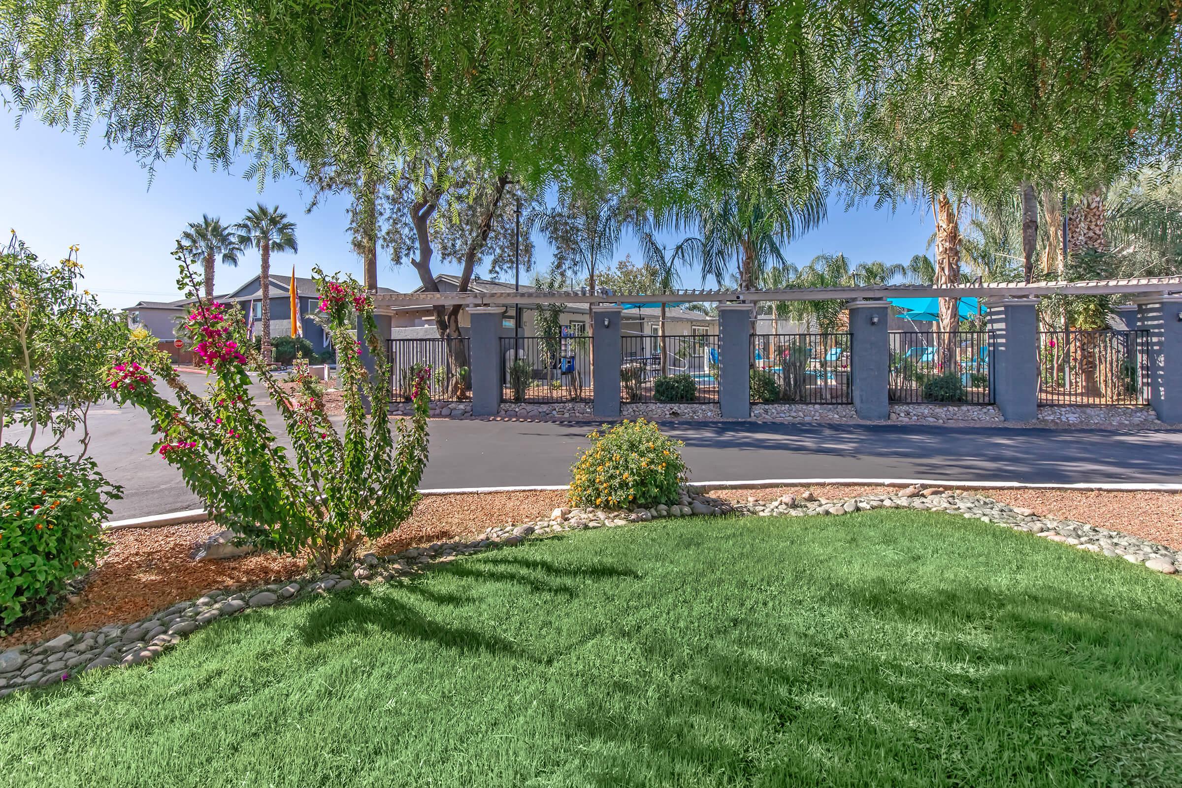 A landscaped view featuring vibrant flowers and green grass in the foreground, with a gated entrance visible in the background. Palm trees and other plants line the area, creating a serene and inviting atmosphere. The sky is clear and sunny, enhancing the overall bright, cheerful setting.