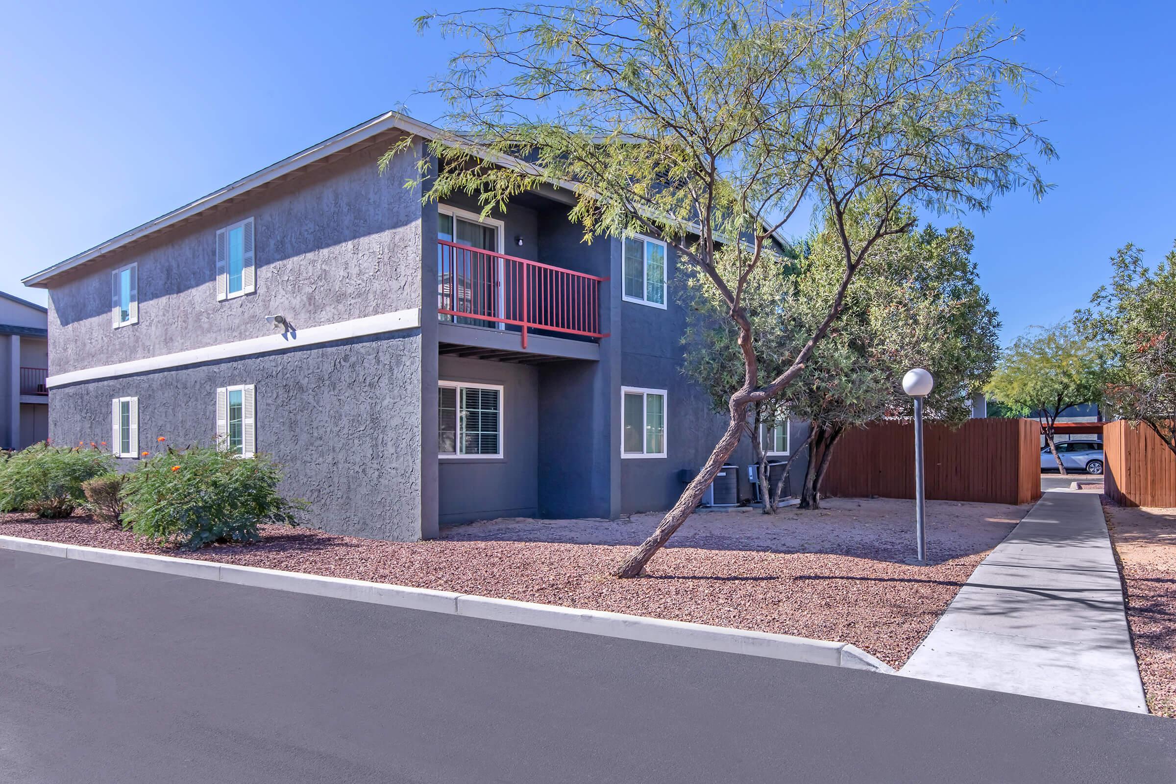 Two-story gray apartment building with a balcony, surrounded by landscaped greenery and pathways. A tree is in the foreground, and a gravel area adds to the outdoor space. Clear blue sky is visible.