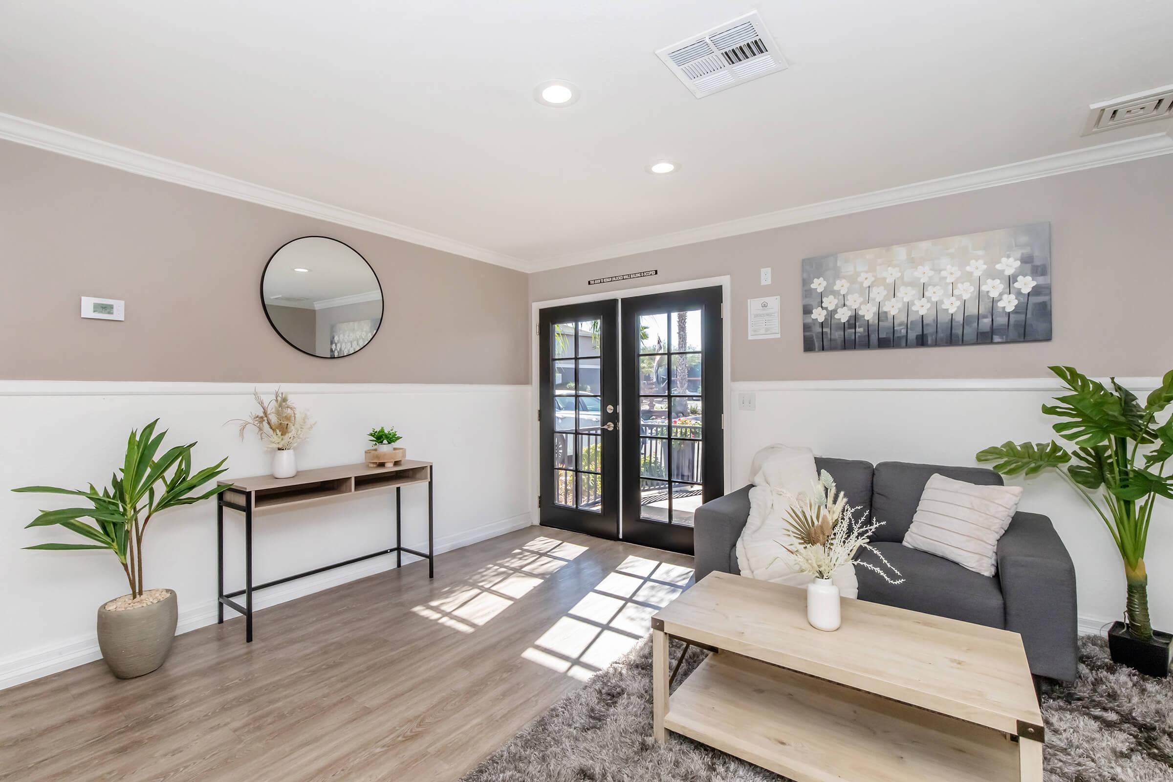 A cozy living room featuring a gray sofa and a wooden coffee table on a plush rug. A potted plant is placed beside a modern console table. Large French doors allow natural light to enter, complementing the neutral-colored walls and decorative mirror. A piece of abstract art hangs above the table, adding a touch of elegance.