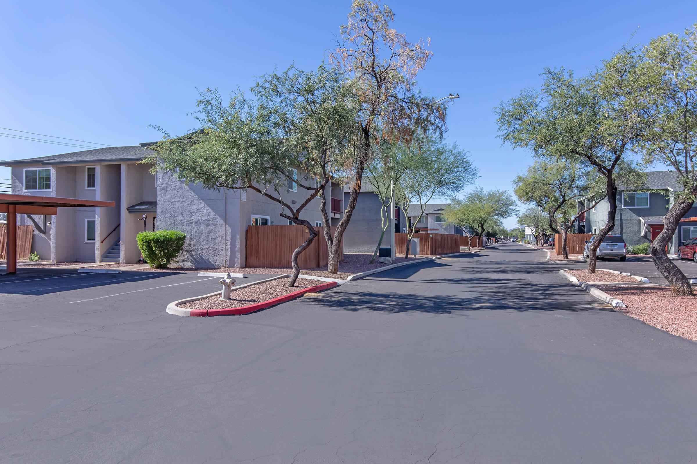 A quiet residential street lined with trees and apartment buildings. The road is paved and has designated parking spaces. There are light shrubs and a clear blue sky overhead, creating a calm atmosphere.