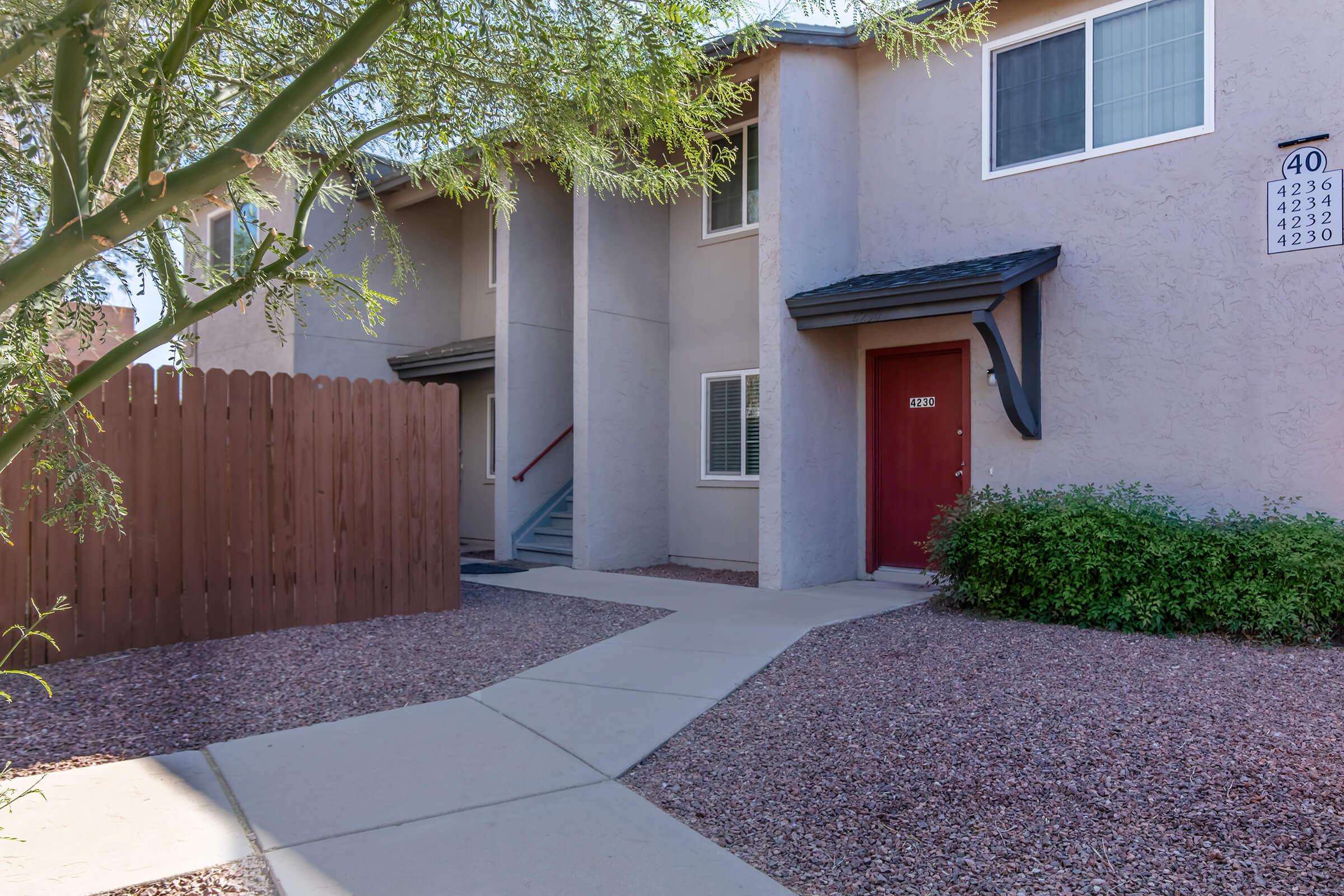 A view of an apartment complex featuring a beige exterior with a red door. A concrete pathway leads to the entrance, surrounded by gravel landscaping and small shrubs. A wooden fence is visible on the left, and a staircase leads to the second floor.