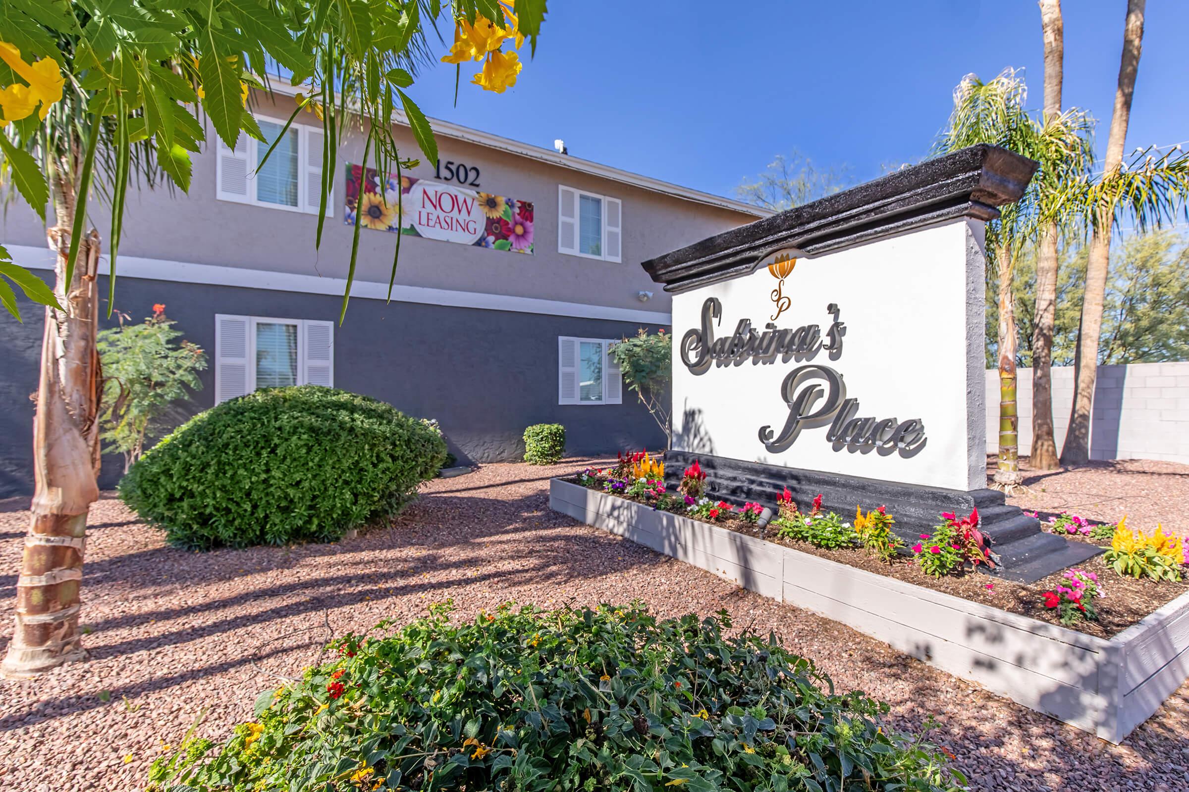 Sign for "Sabrina's Place" with lush landscaping, colorful flowers, and a banner stating "Now Leasing." The building in the background is two stories with a gray exterior.