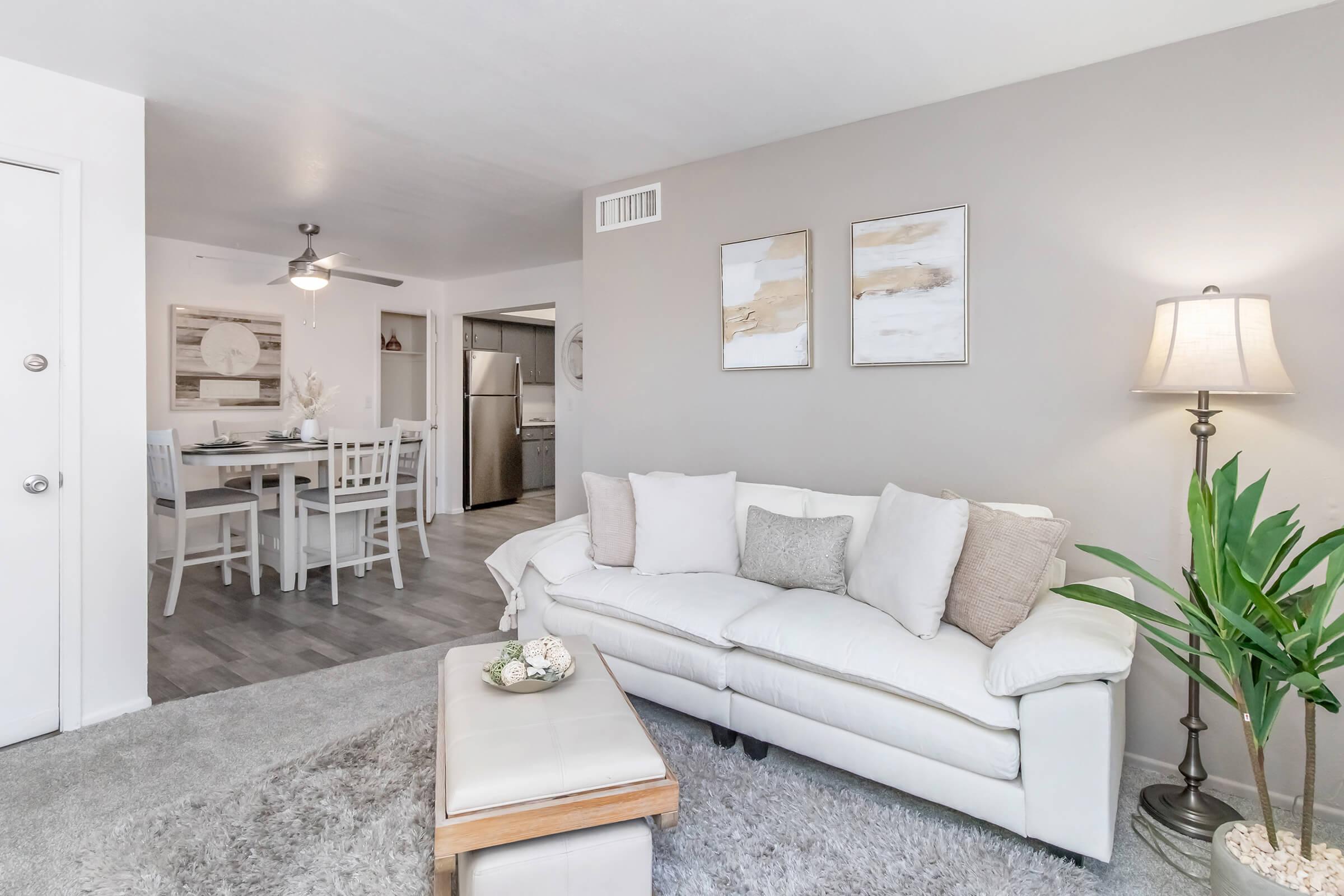 A modern living room featuring a white couch with decorative pillows and a small coffee table. A floor lamp illuminates the space, and a green potted plant adds a touch of nature. In the background, there is a dining area with a table and chairs, and a glimpse of a kitchen. The walls are painted in a soft gray tone.