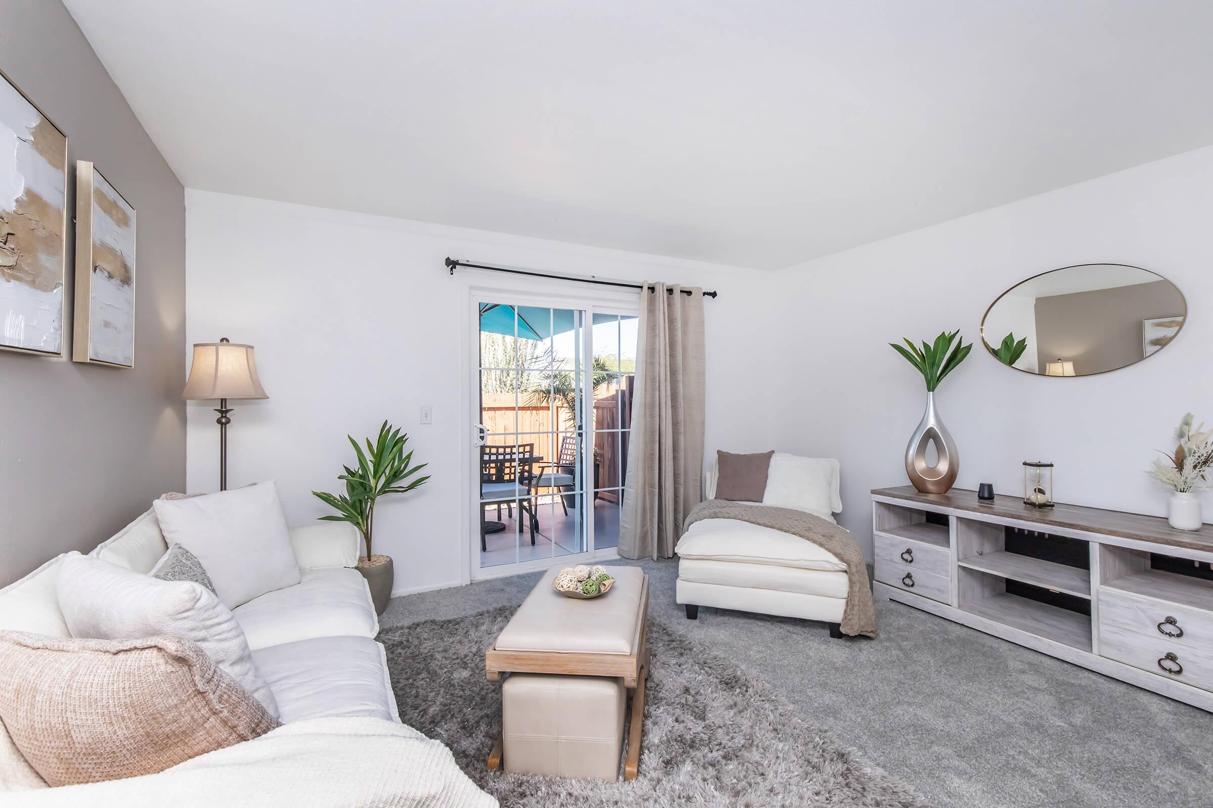 A cozy living room featuring a white sofa, a chaise lounge, and a wooden coffee table on a soft rug. The walls are light-colored with decorative artwork, and a floor lamp adds warmth. A sliding glass door leads to an outdoor area with patio furniture, framed by curtains. Green plants provide a touch of nature.
