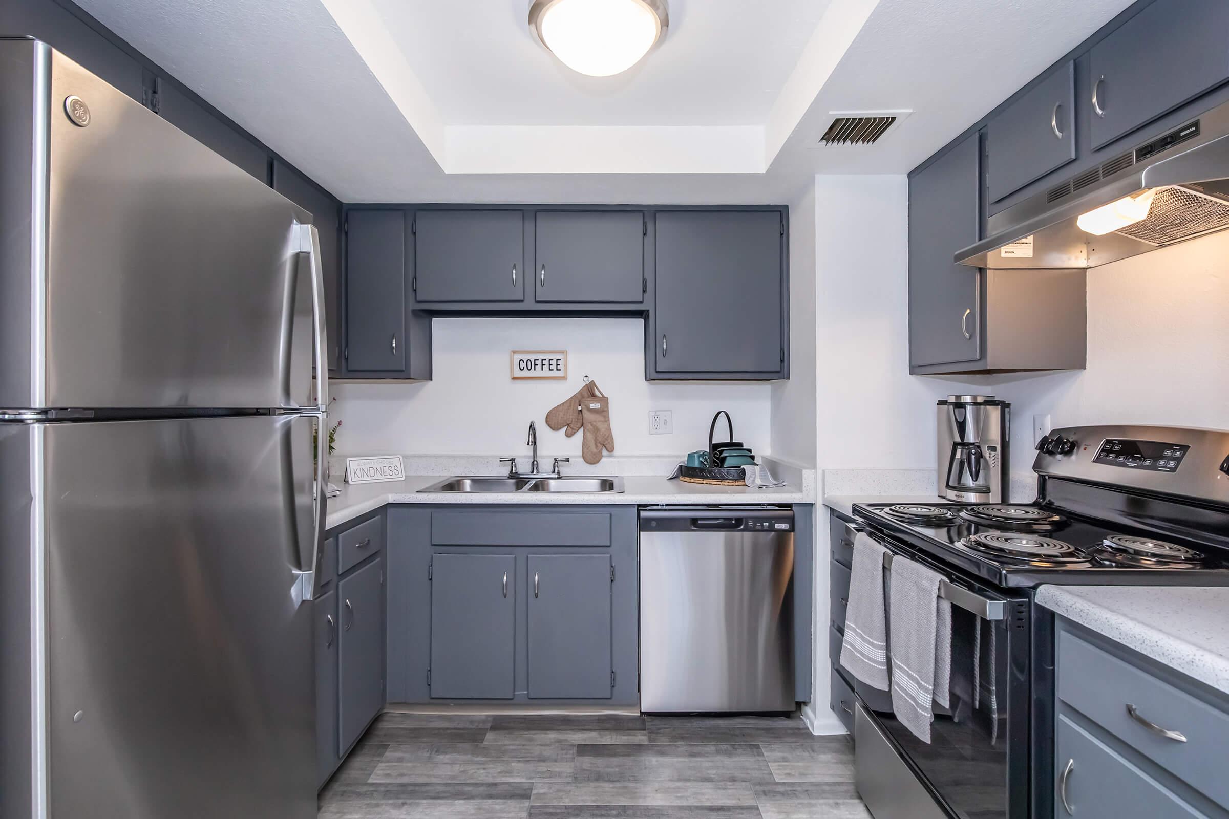 Modern kitchen featuring gray cabinetry, stainless steel appliances, and a light-colored countertop. Includes a refrigerator, dishwasher, and stove, with a coffee-themed decoration on the wall. The floor is tiled with a light wood appearance, providing a bright and contemporary atmosphere.