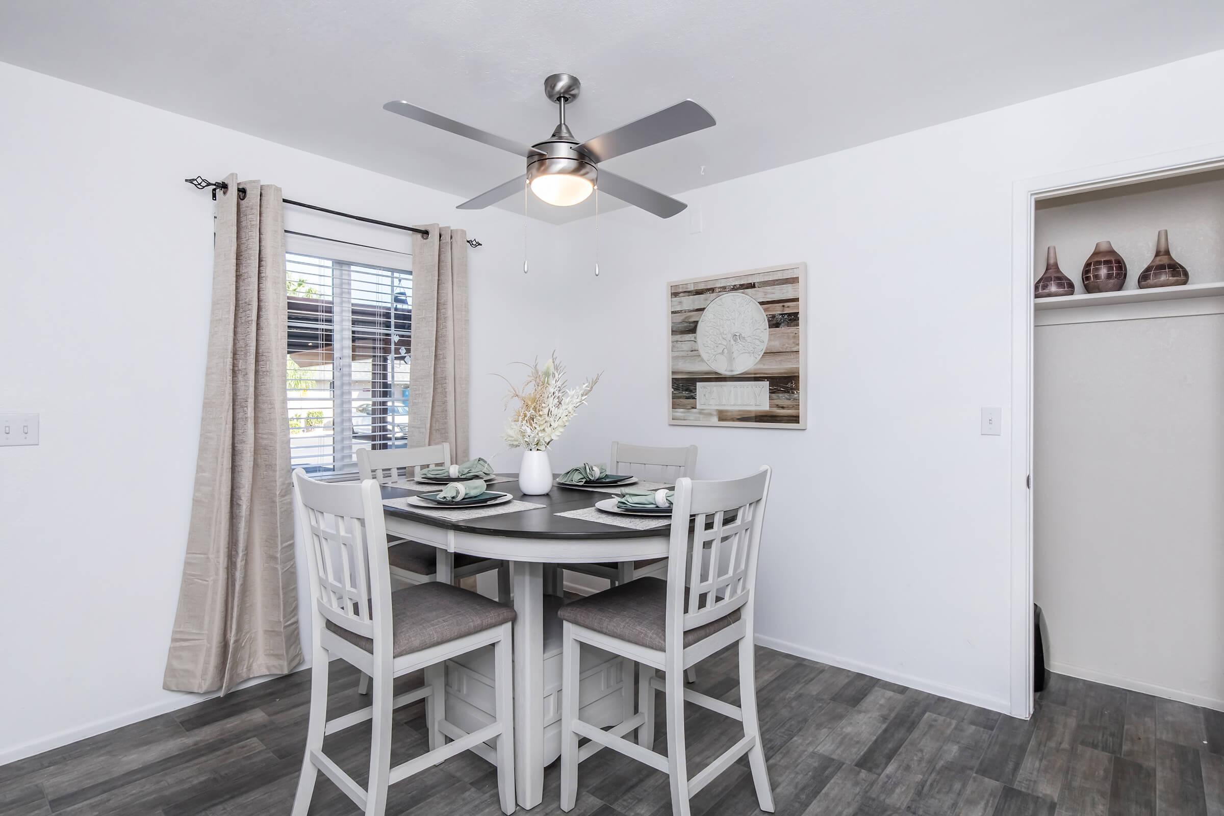 A bright dining area featuring a round table with four chairs, a modern ceiling fan, and large windows with light curtains. A decorative wall art piece and a closet add to the ambiance, creating a cozy and inviting atmosphere for dining or gathering.