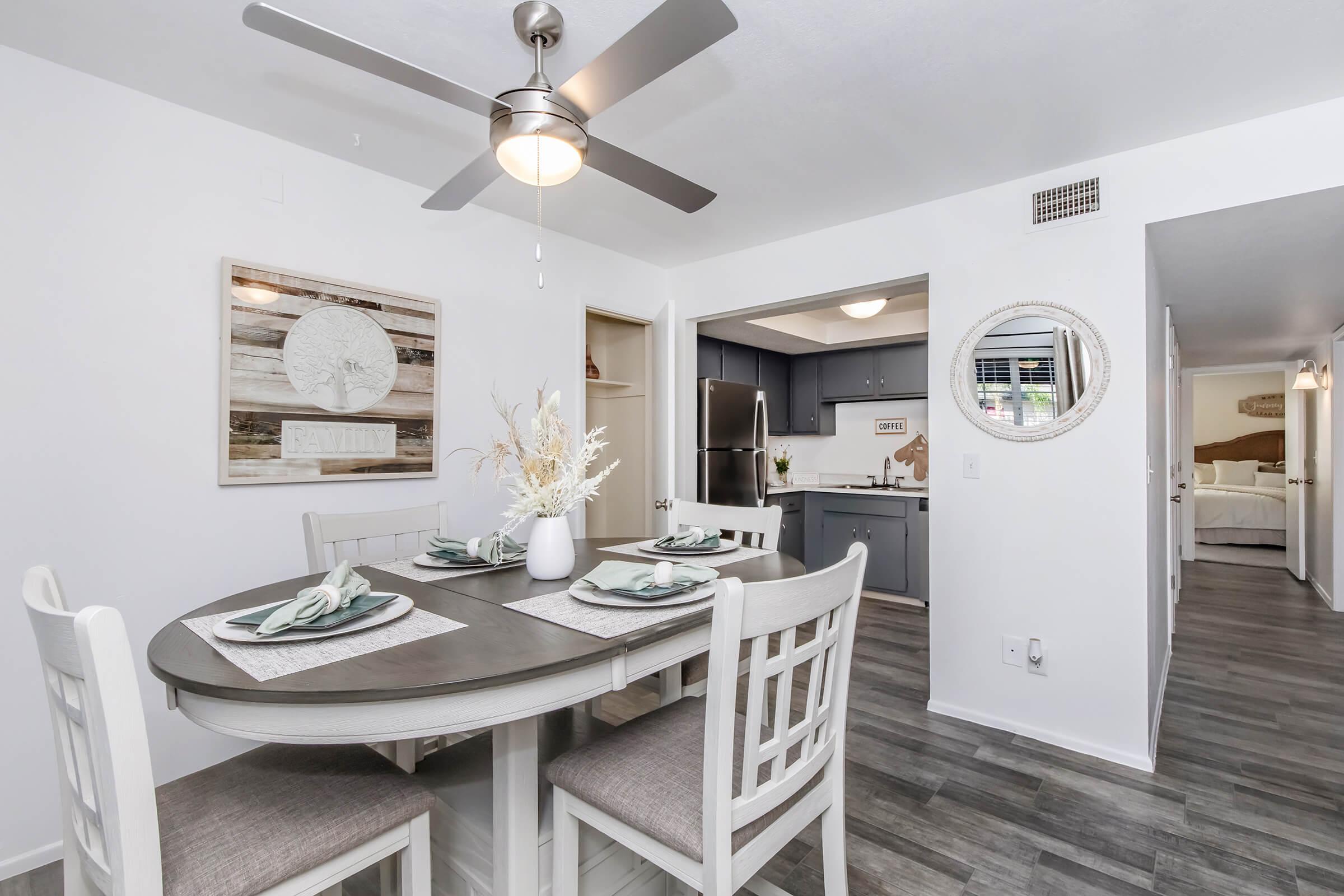 A modern dining area featuring a round table set for four with decorative placemats and a vase with dried flowers. A ceiling fan is above, and there’s a framed wall art piece nearby. The background shows a kitchen with stainless steel appliances and an adjacent room visible through an open door.