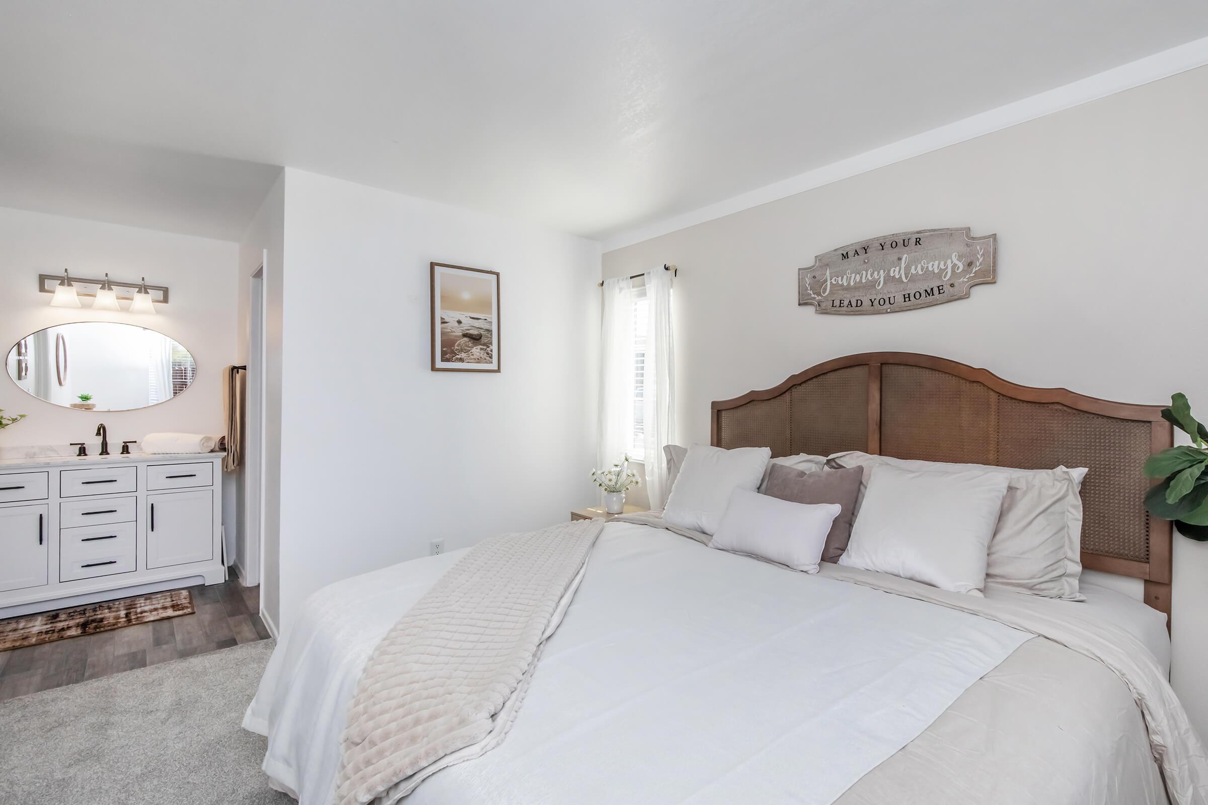 Bright and airy bedroom featuring a large bed with neutral-colored bedding and decorative pillows. A wooden headboard adds warmth to the space. In the background, a bathroom with a mirror and vanity is visible. Natural light streams through the windows, creating a cozy, inviting atmosphere.