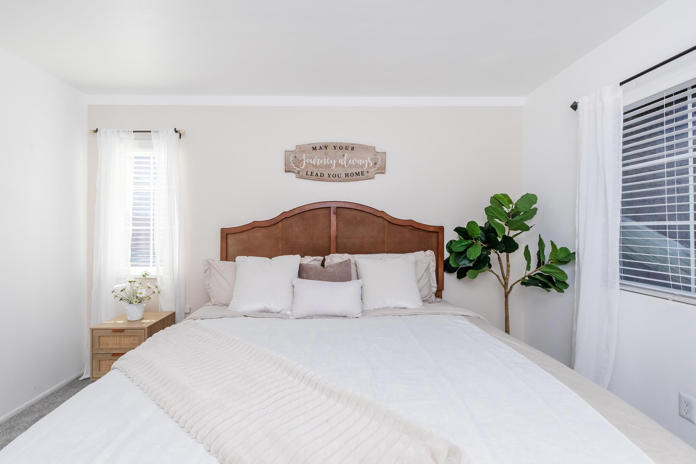 A cozy bedroom featuring a large bed with neutral bedding, a wooden headboard, and decorative pillows. There's a bedside table with a small plant and a vase of flowers. Sunlight filters through sheer white curtains, and a wall sign above the bed reads "May your dreams lead you home."