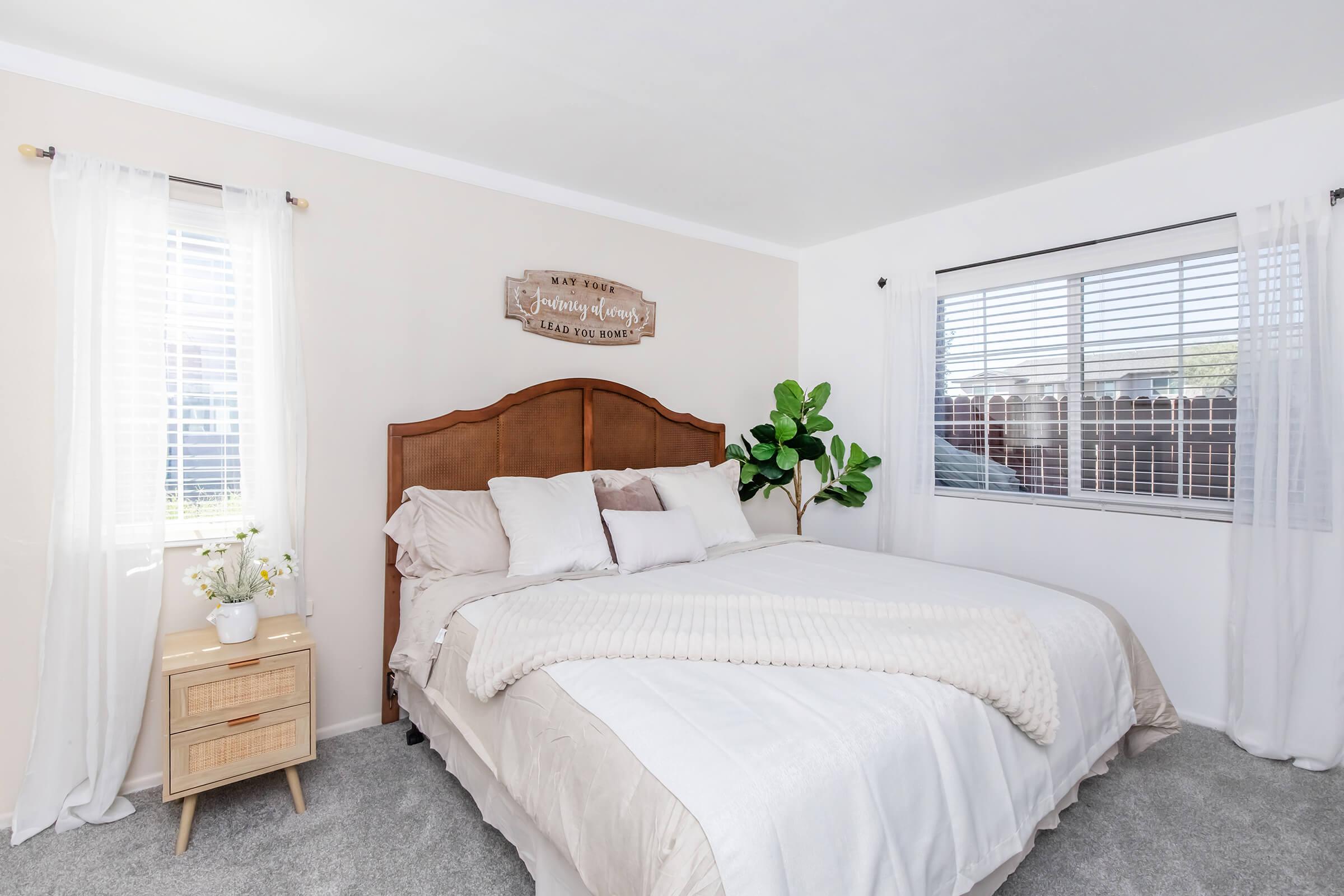 Cozy bedroom featuring a large bed with soft linens, decorative pillows, and a neutral color scheme. A wooden sign hangs above the bed, and there's a decorative plant in the corner. Sheer curtains allow natural light through the windows, creating a warm and inviting atmosphere. A nightstand with a small vase of flowers adds a personal touch.