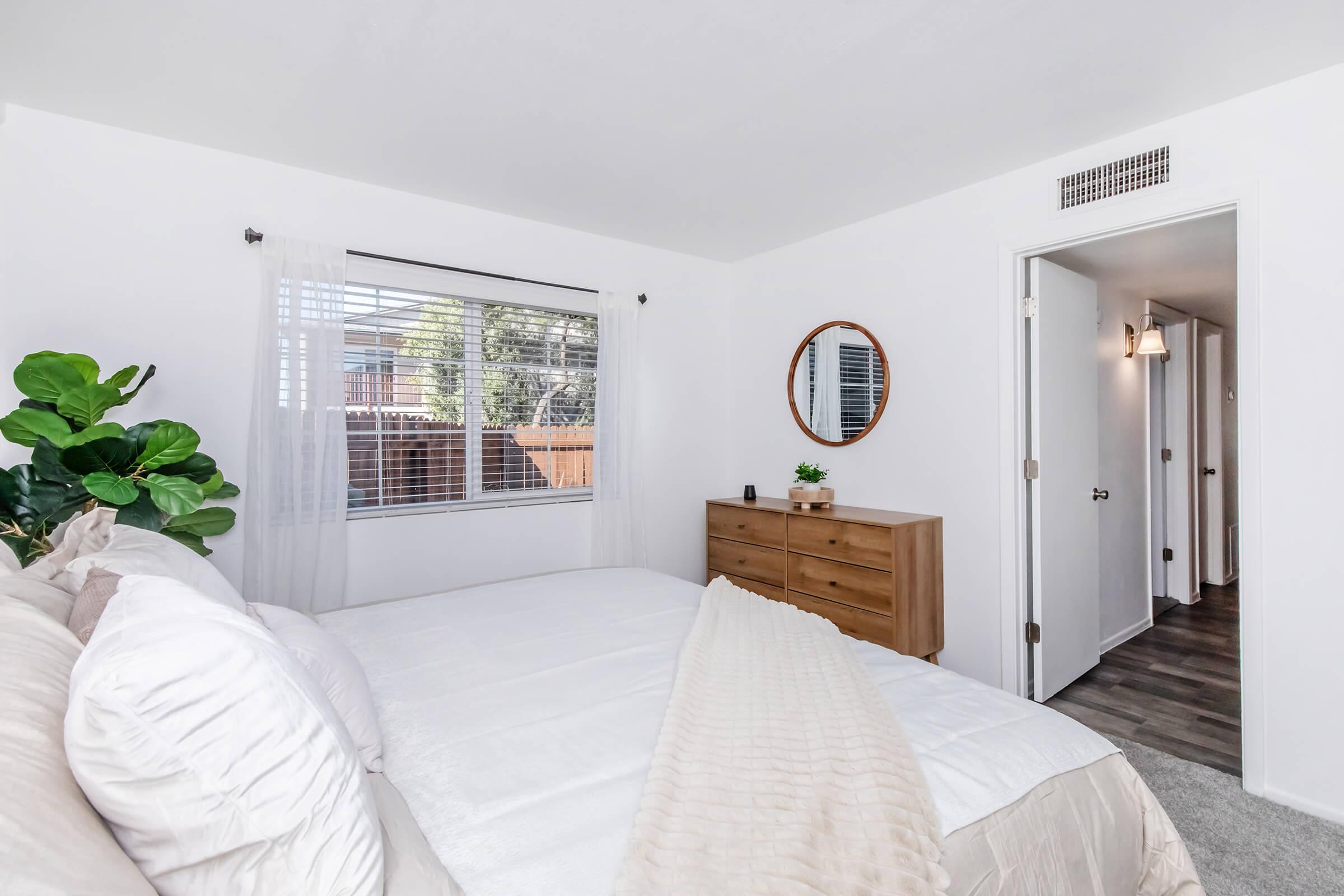 A cozy bedroom featuring a double bed with white linens, a wooden dresser, and a round mirror. Natural light filters through a window adorned with sheer curtains. A green plant adds a touch of nature, while a hallway is visible in the background, leading to additional spaces.