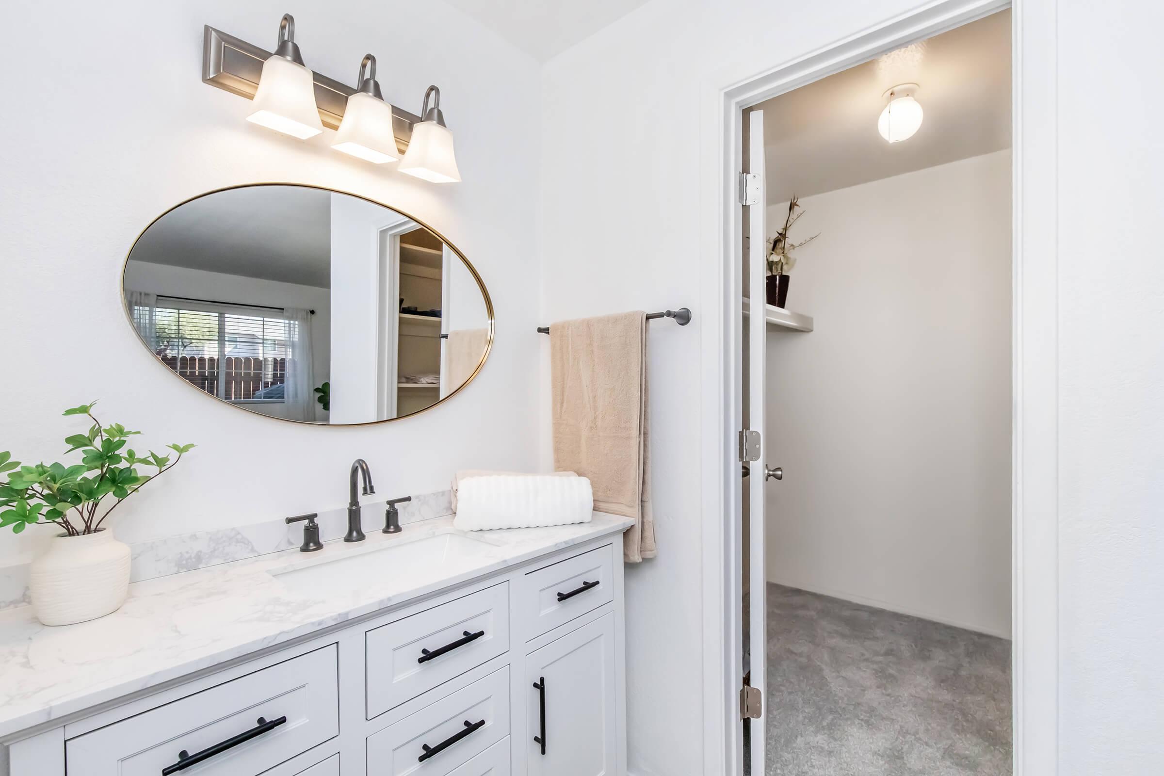 Modern bathroom with a round mirror above a white marble countertop. The countertop features a sleek faucet and a white towel. Soft lighting is provided by three sconces above the mirror. A doorway leads to a carpeted room with minimal decor. A potted plant adds a touch of greenery to the space.
