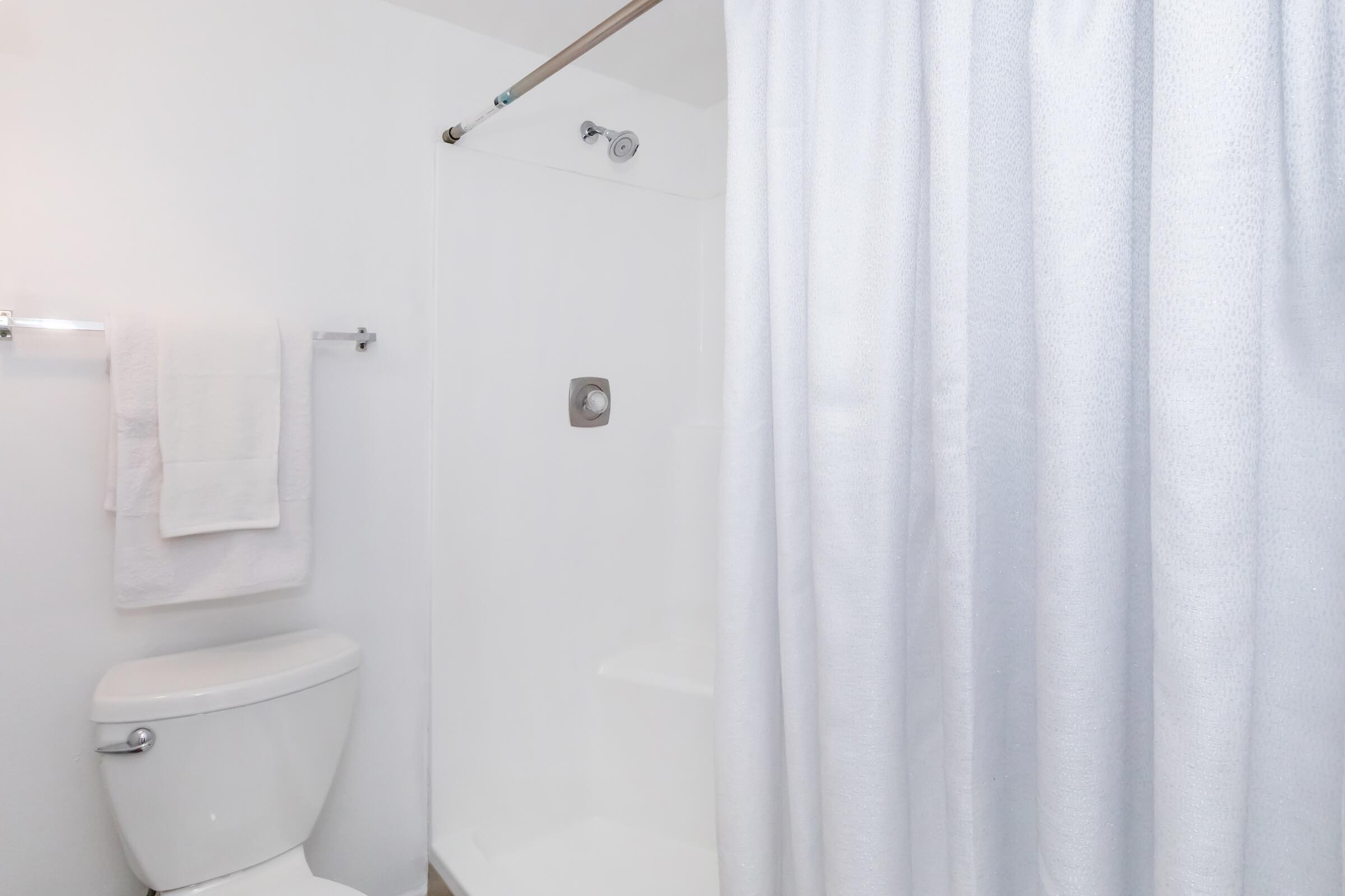 A clean bathroom scene featuring a white shower curtain, a shower stall with a silver faucet, and a toilet. Soft white towels hang on the wall, creating a bright and tidy atmosphere. The walls are a light color, contributing to a fresh and spacious feel.