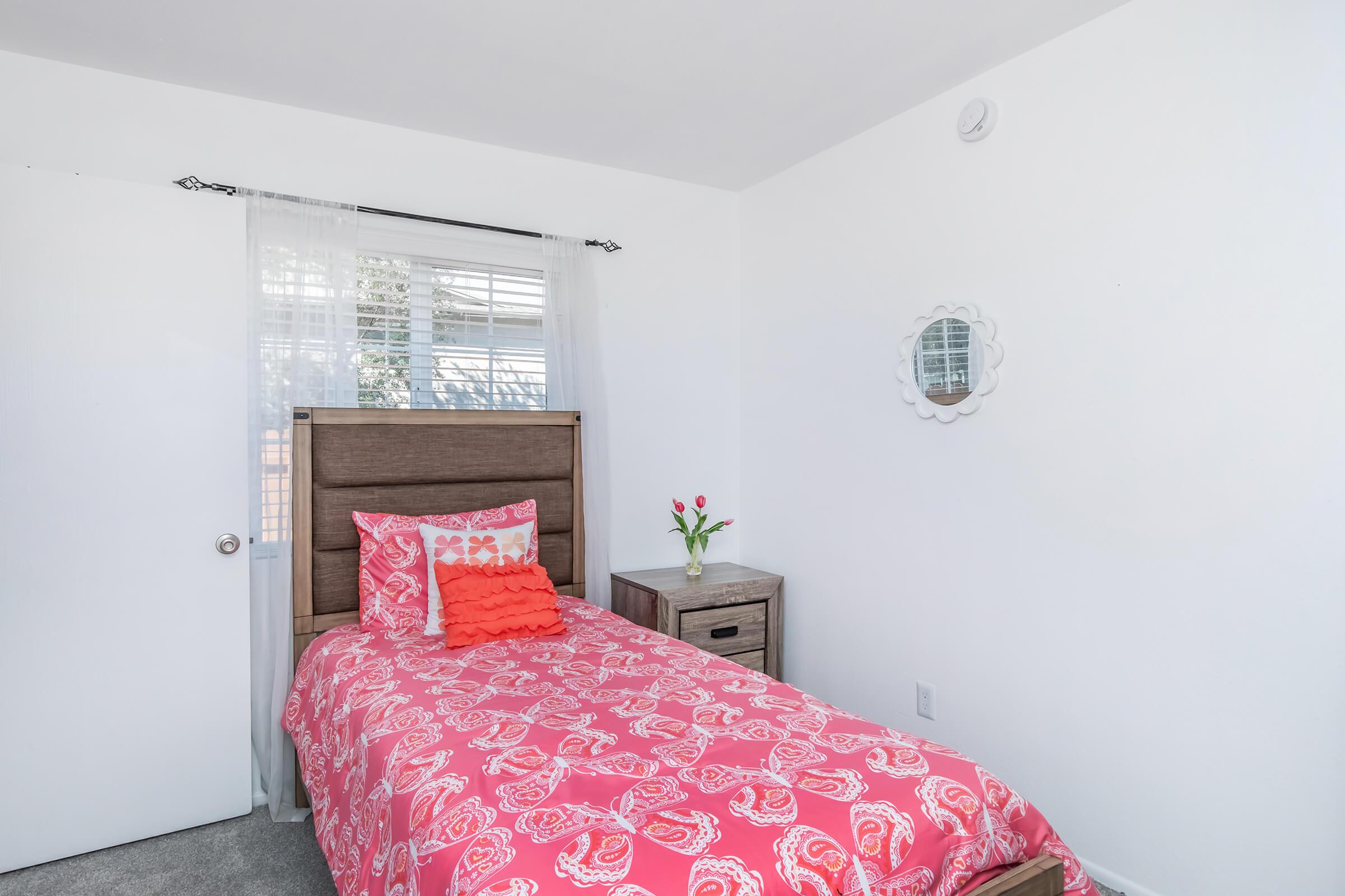 A bright and tidy bedroom featuring a single bed with a pink patterned comforter and decorative pillows. The room has white walls and a window draped with sheer curtains, allowing natural light. A bedside table holds a vase with tulips, and a round mirror is mounted on the wall.